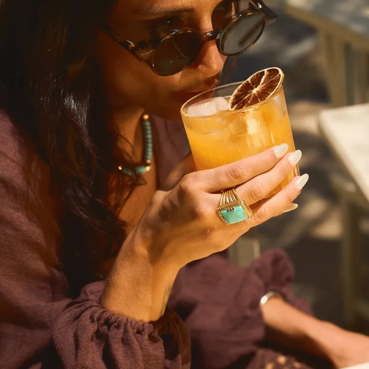 Woman wearing sunglasses and a turquoise ring holding a glass of iced orange cocktail garnished with a dried citrus slice.
