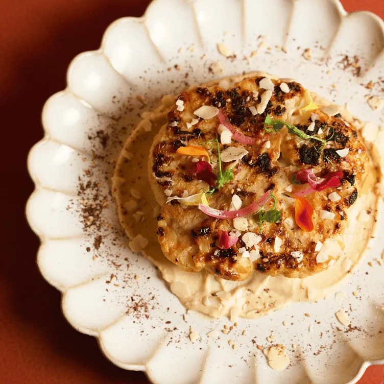Grilled food served on a white scalloped plate with creamy sauce, garnished with sliced almonds, herbs, and edible flower petals.