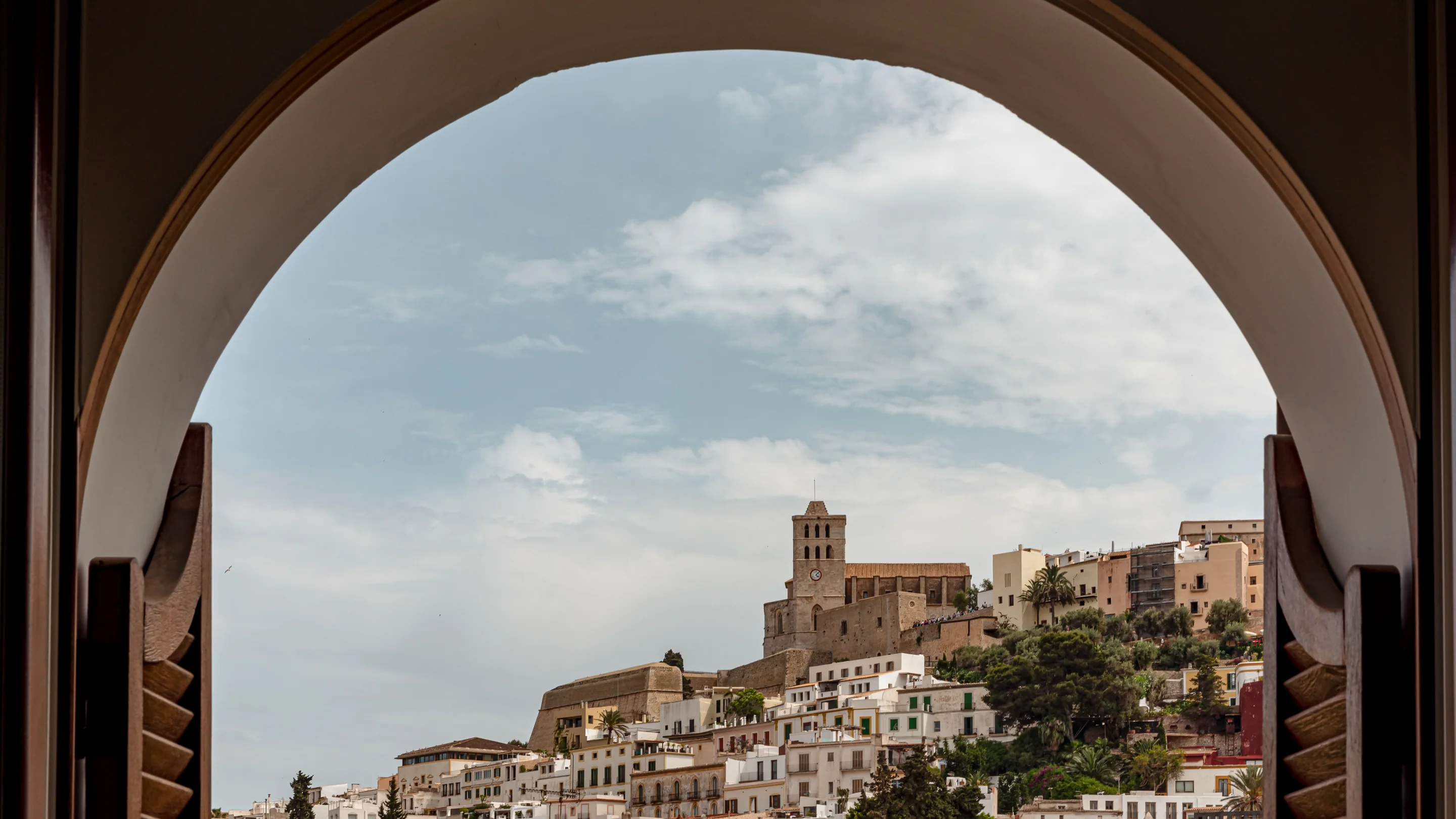 View of a historic town with white buildings and a clock tower church seen through a large arched window frame.