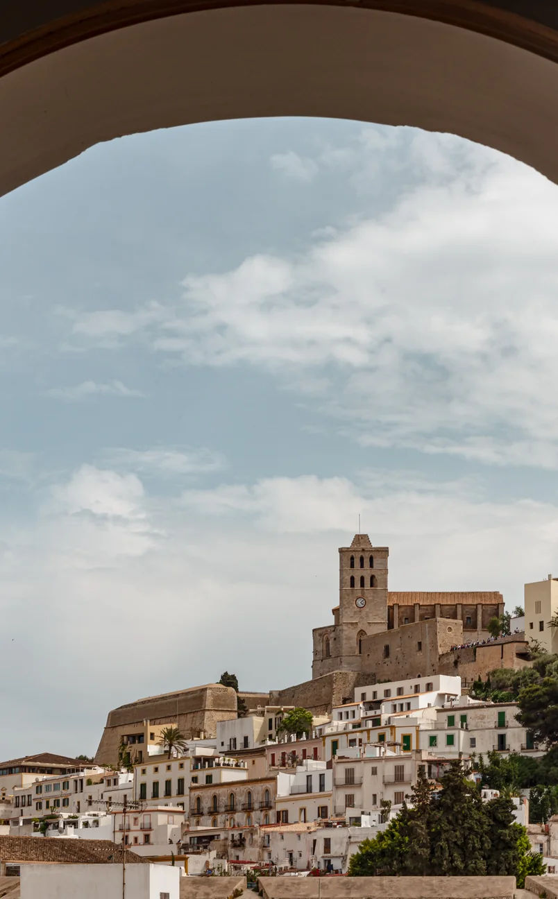 View of a historic fortress and church tower overlooking white buildings under a partly cloudy sky, framed by a large arch.
