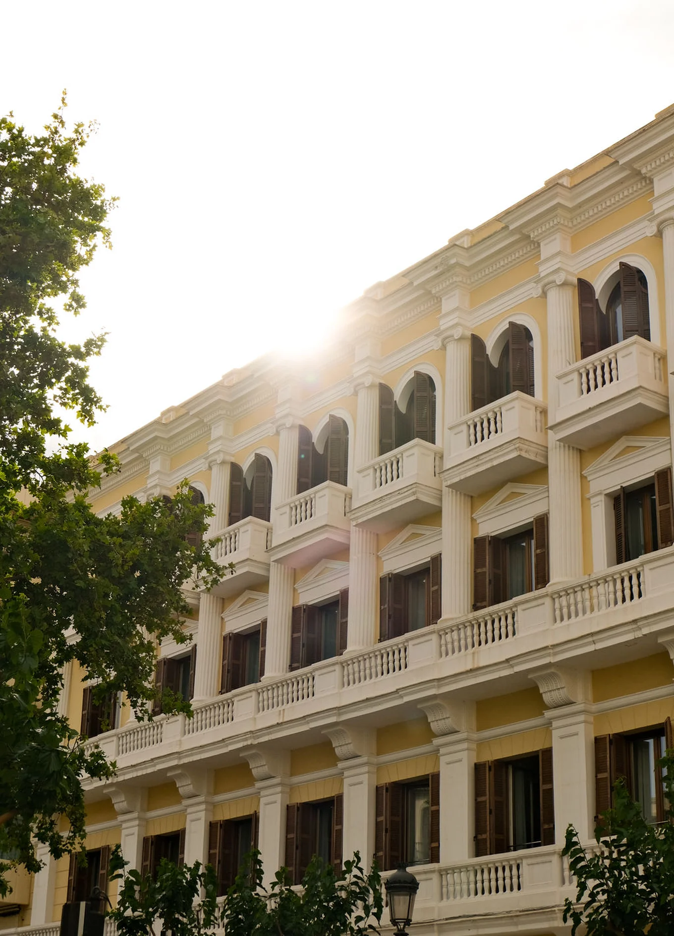 Sunlight shining over the yellow facade of a classic-style building with arched windows, wooden shutters, and balconies.
