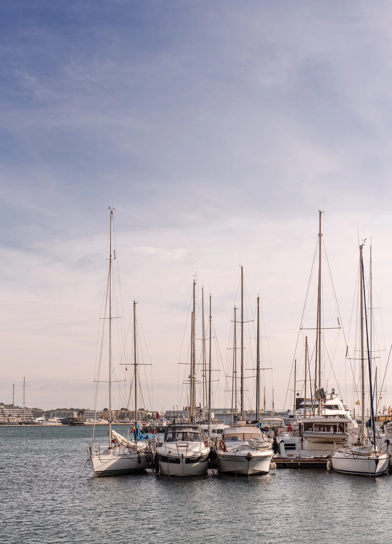Several sailboats and yachts docked side by side in a calm marina under a partly cloudy sky.