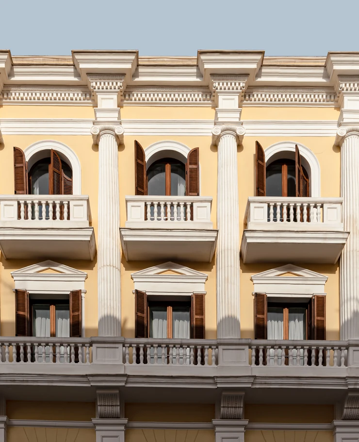 Facade of a classical yellow building with white columns, balconies, and brown wooden shutters on arched windows.