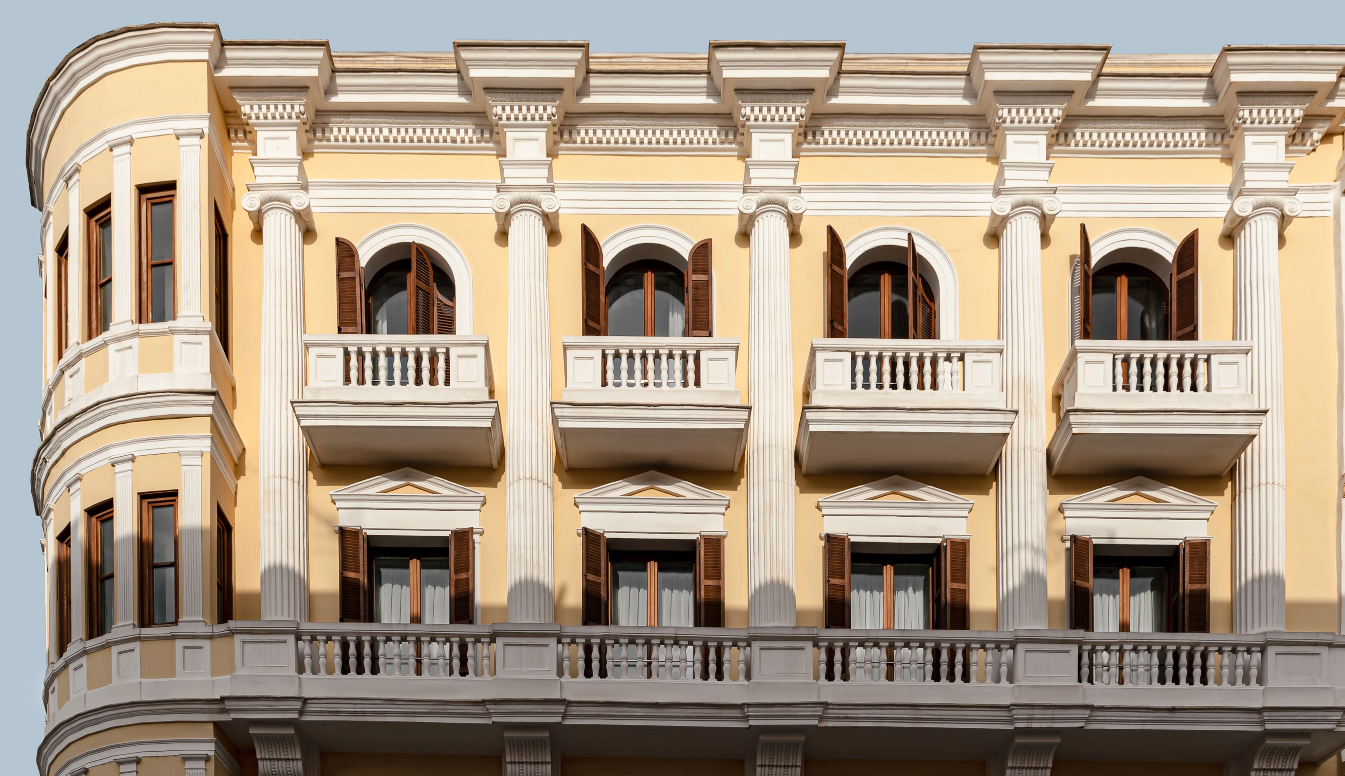 Yellow building façade with white classical columns and wooden shutters on arched windows and balconies.