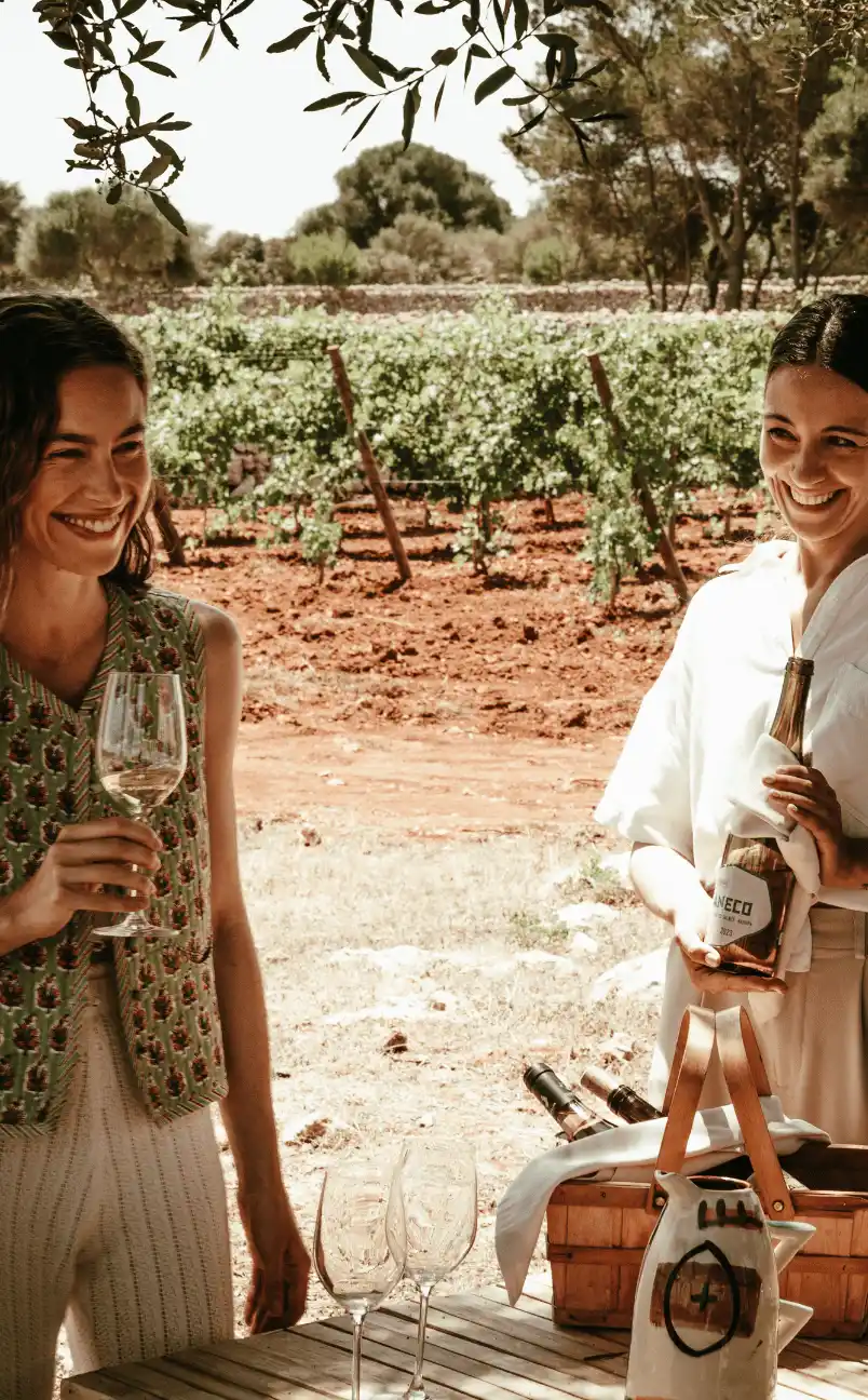 Two smiling women enjoying wine tasting outdoors near a vineyard, holding a wine glass and a wine bottle.