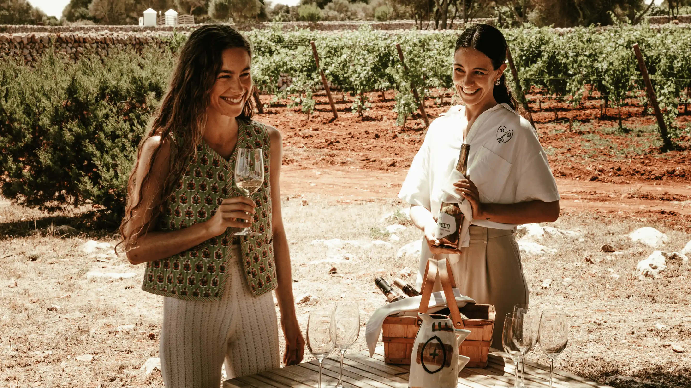 Two women smiling and enjoying wine outdoors near vineyard, one holding a wine bottle and the other a glass.