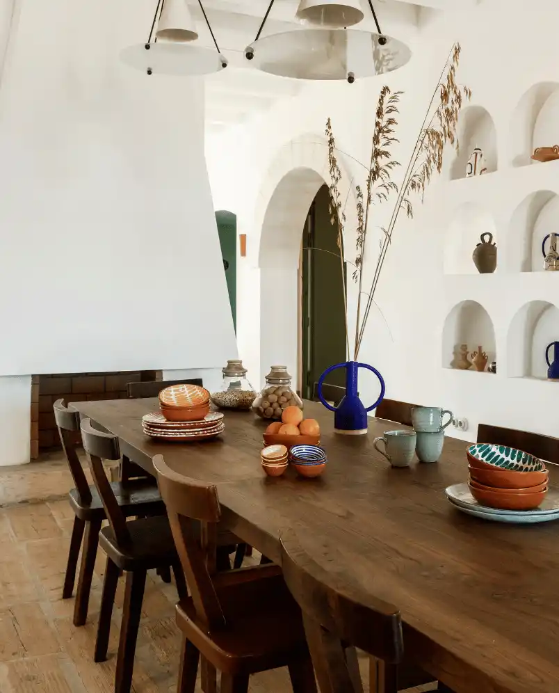 Long wooden dining table with six chairs set with ceramic bowls, plates, cups, and a blue vase holding dried grasses in a bright Mediterranean-style room.