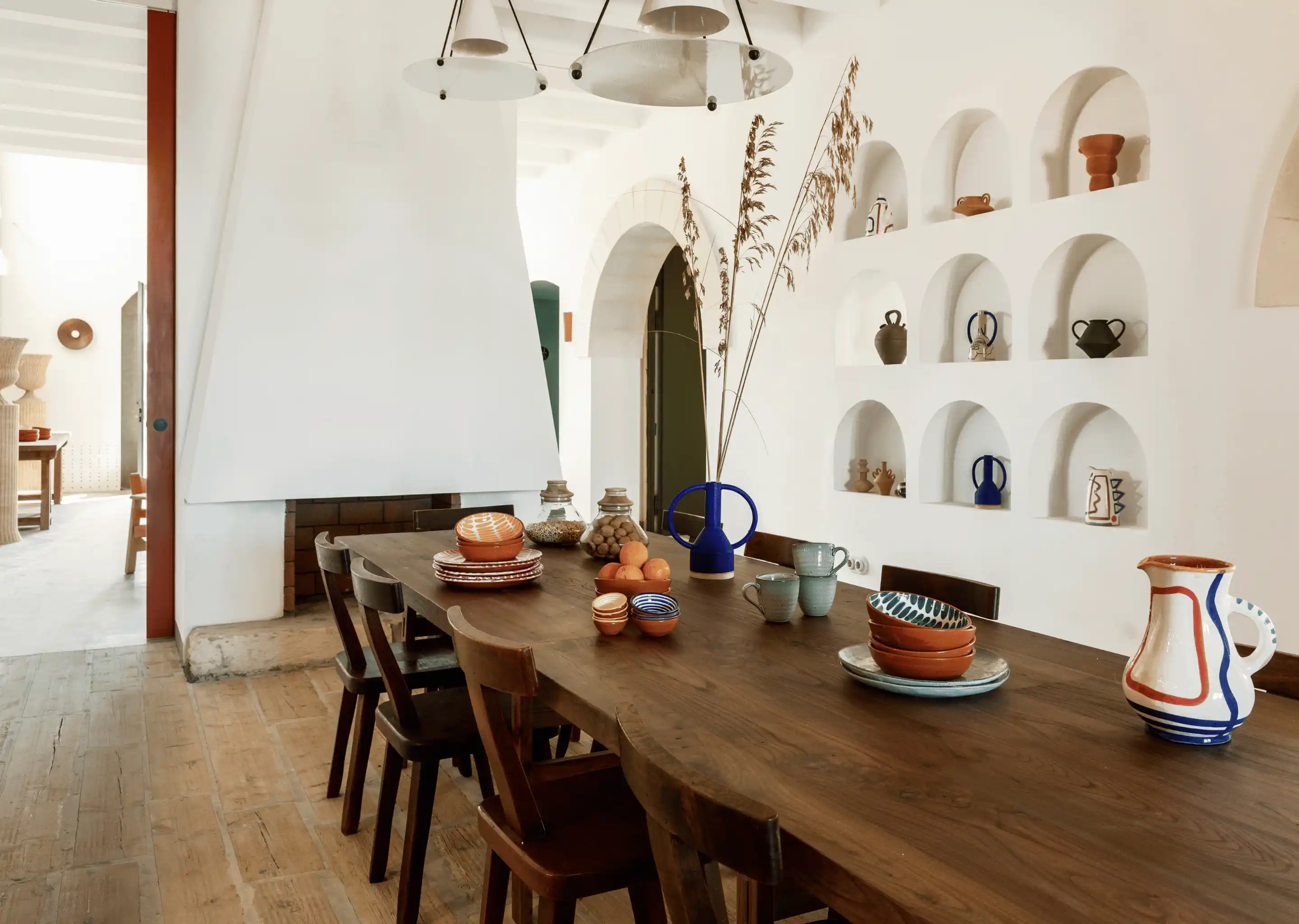 Dining table with wooden chairs, ceramic dishes, and decorative vases in a bright room with arched wall niches.