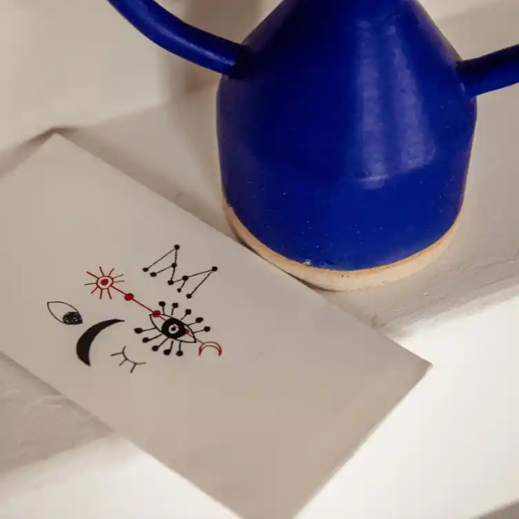 Blue ceramic teapot beside a white card with abstract eye and geometric line art in black and red.