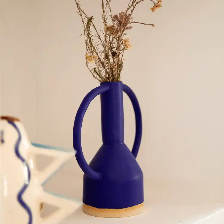 Blue vase with large round handles holding dried flowers on a white surface.