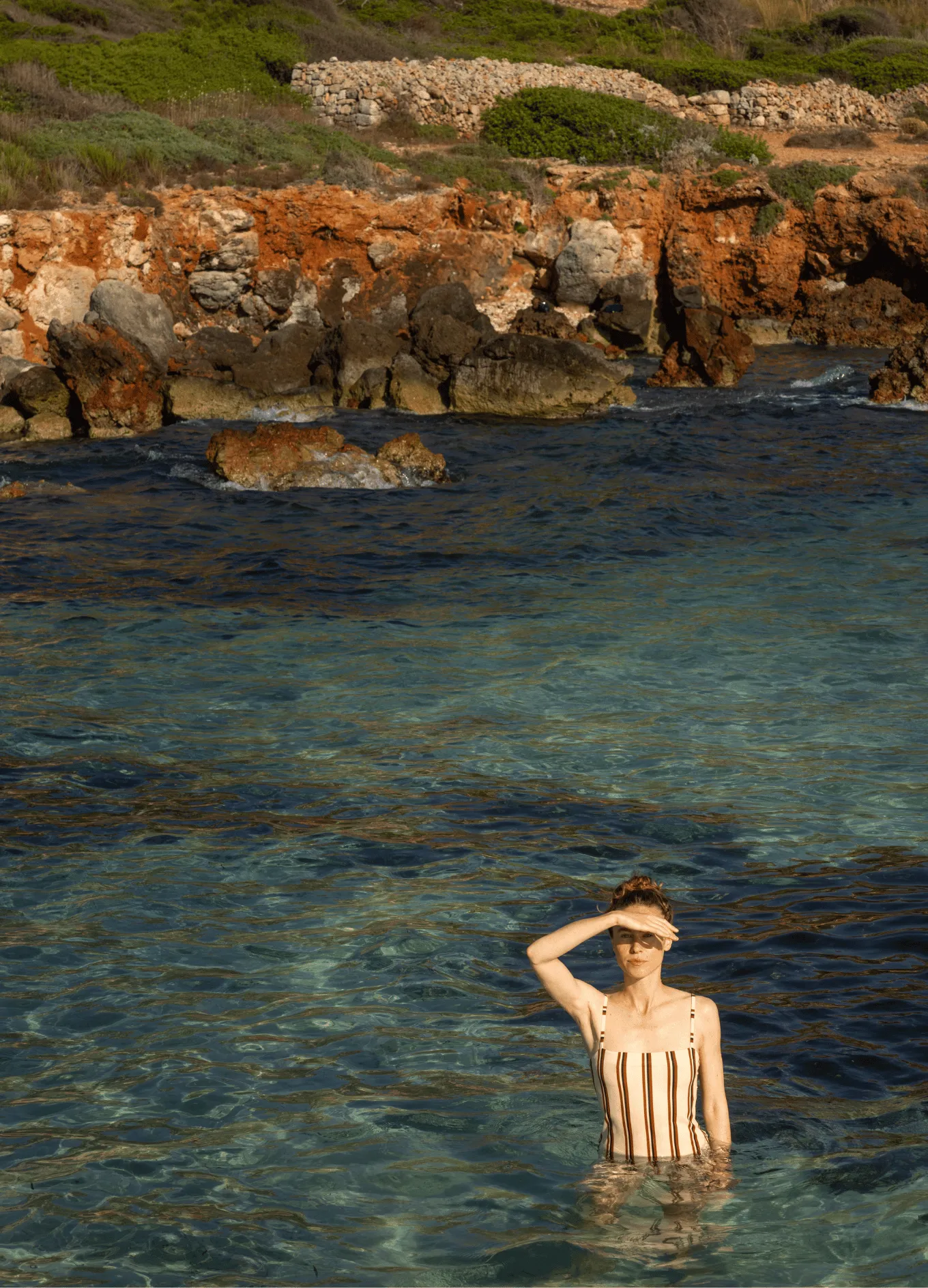 Woman in striped swimsuit standing in clear blue sea near rocky shore with greenery.