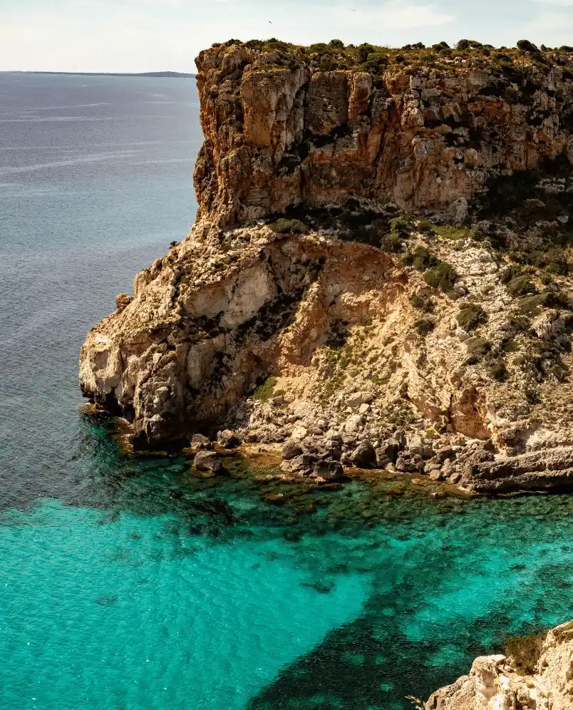 Rocky cliff with sparse vegetation overlooking clear turquoise sea water.