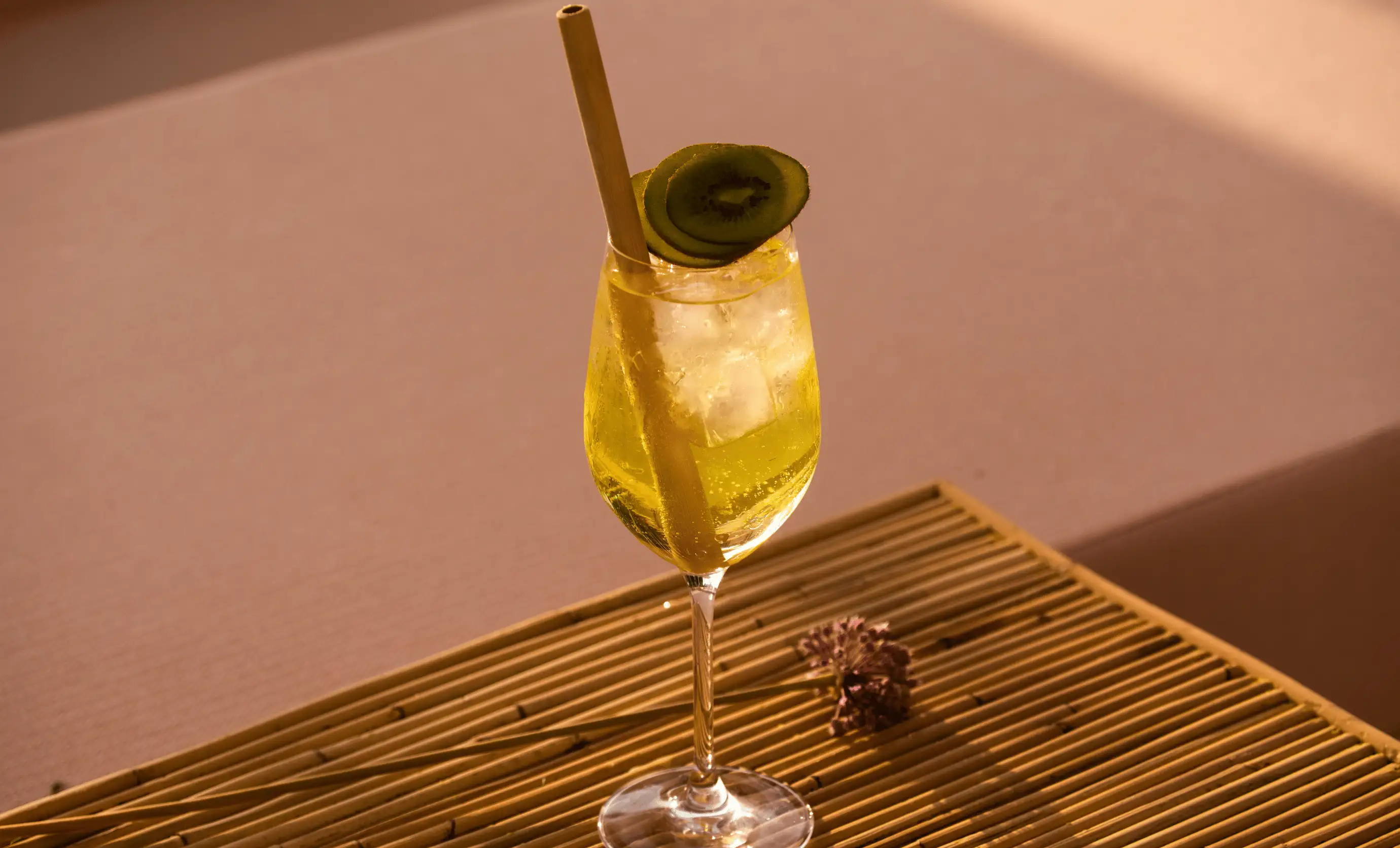 Yellow cocktail with ice cubes, garnished with kiwi slices and a bamboo straw on a bamboo surface.