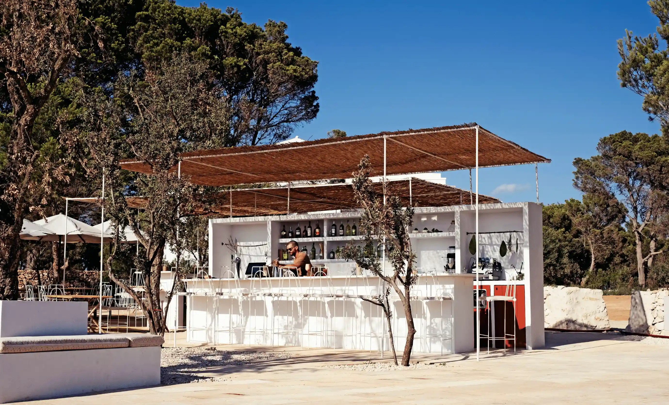 Outdoor hotel bar in Menorca with a man behind the counter, surrounded by tall white chairs under wooden canopy shades and pine trees.