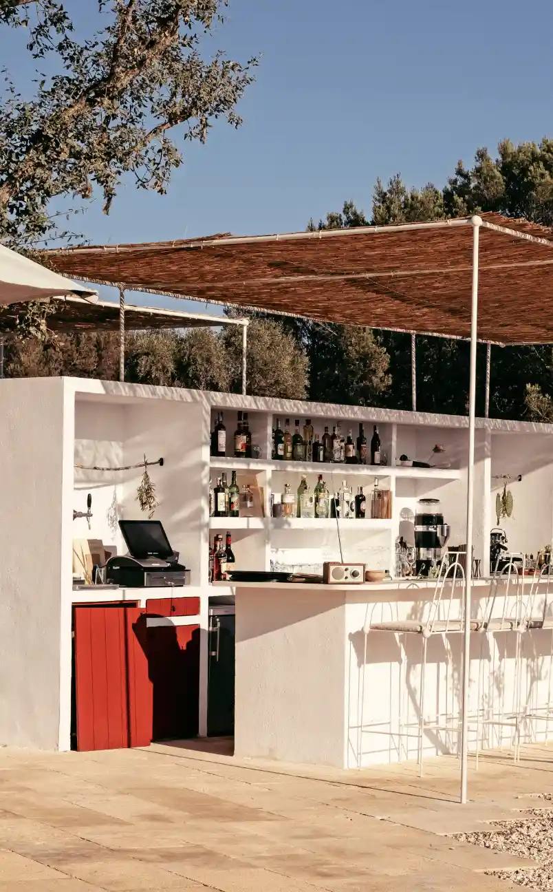 Outdoor bar area with white walls and shelves stocked with bottles, a wooden canopy overhead, and white metal bar stools.