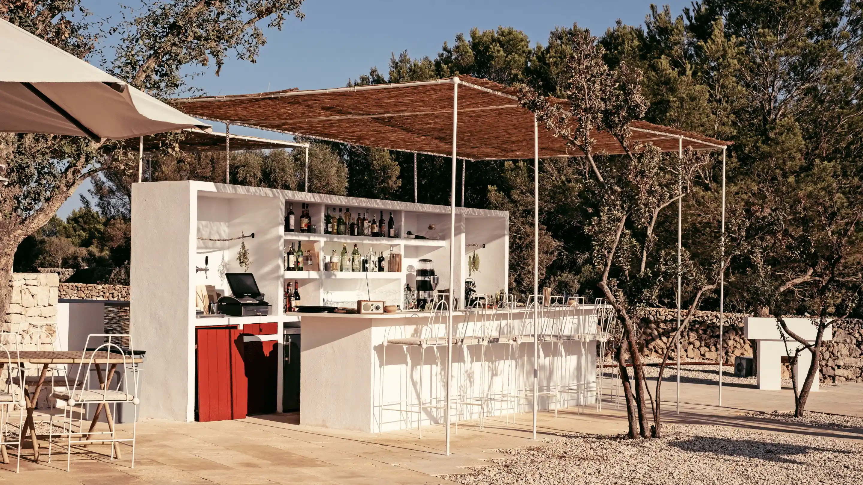 Outdoor bar with white walls, red cabinet doors, white metal bar stools, and a woven canopy roof, surrounded by trees and stone walls.