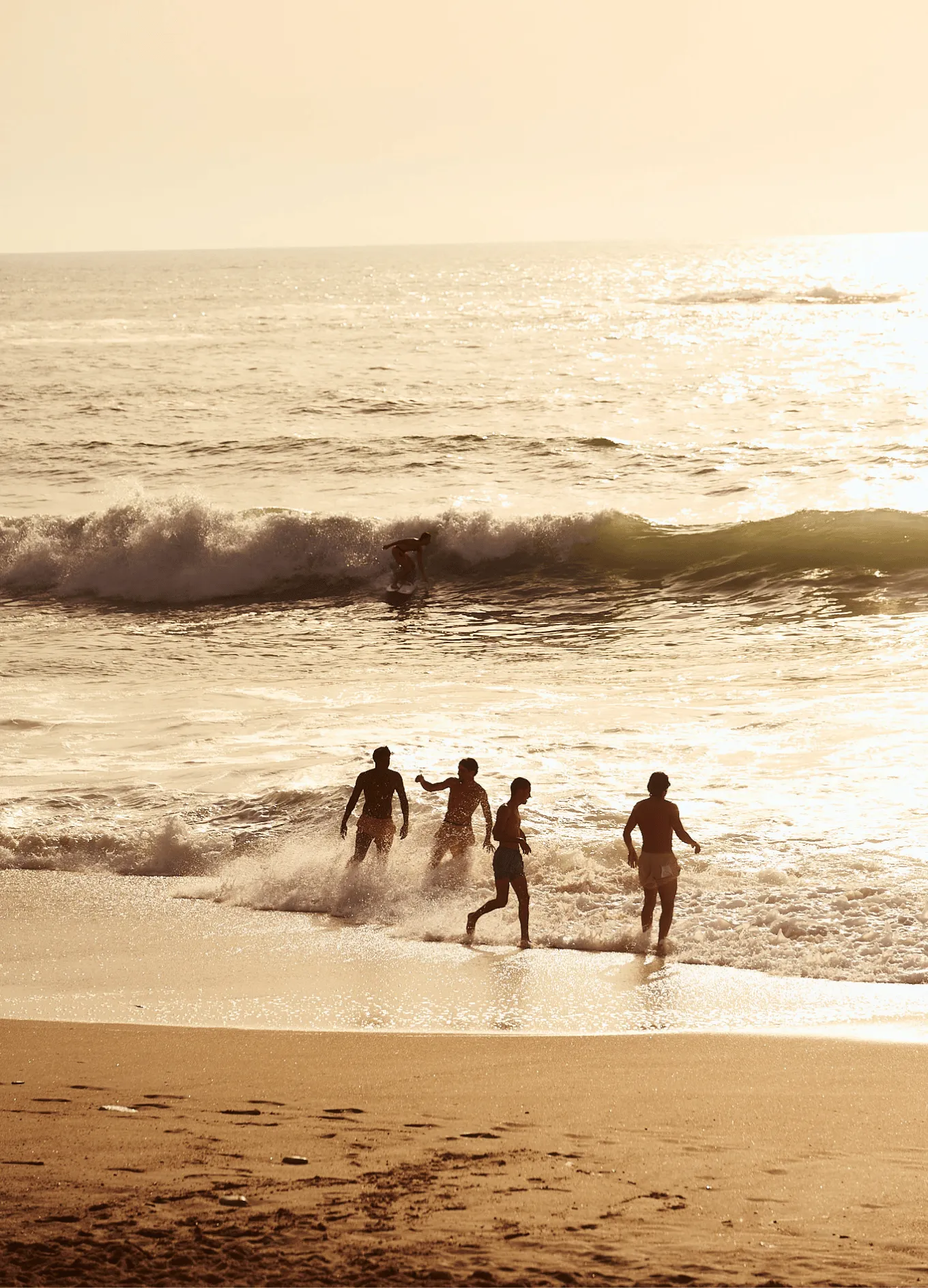 Four people playing near the shoreline and a surfer riding a wave in the sea during sunset.