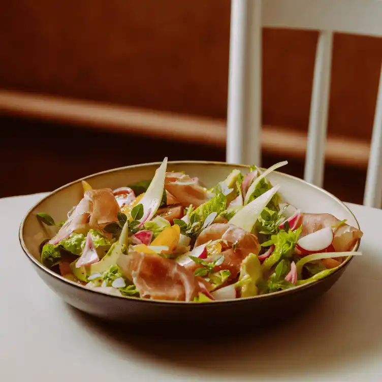 Fresh salad with lettuce, radishes, yellow bell peppers, and slices of cured ham in a shallow bowl on a white table.