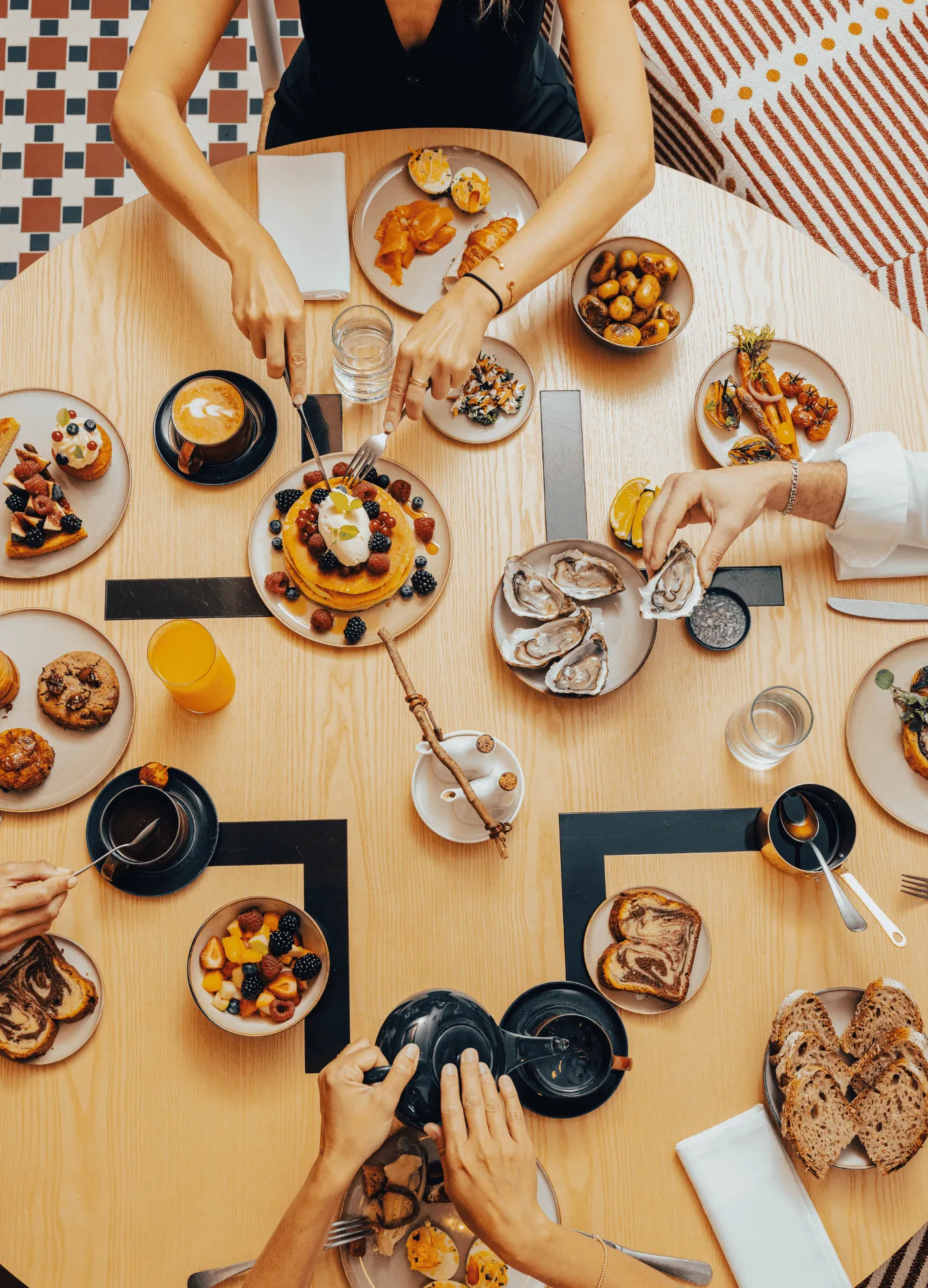 Top view of a table with people enjoying a diverse brunch including pancakes with berries, oysters, fruit bowl, pastries, coffee, and juice.