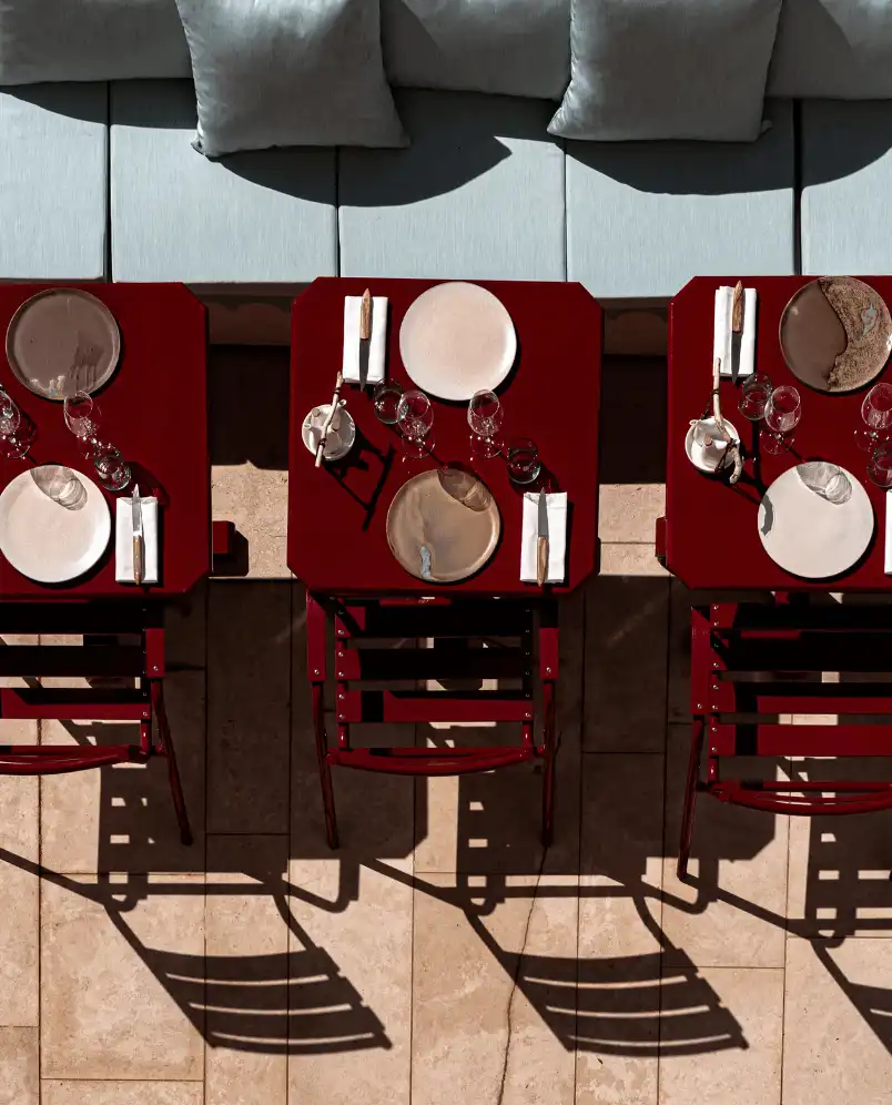 Overhead view of three dark red tables with place settings, glassware, and cutlery next to a grey cushioned bench, casting chair shadows on tiled floor.