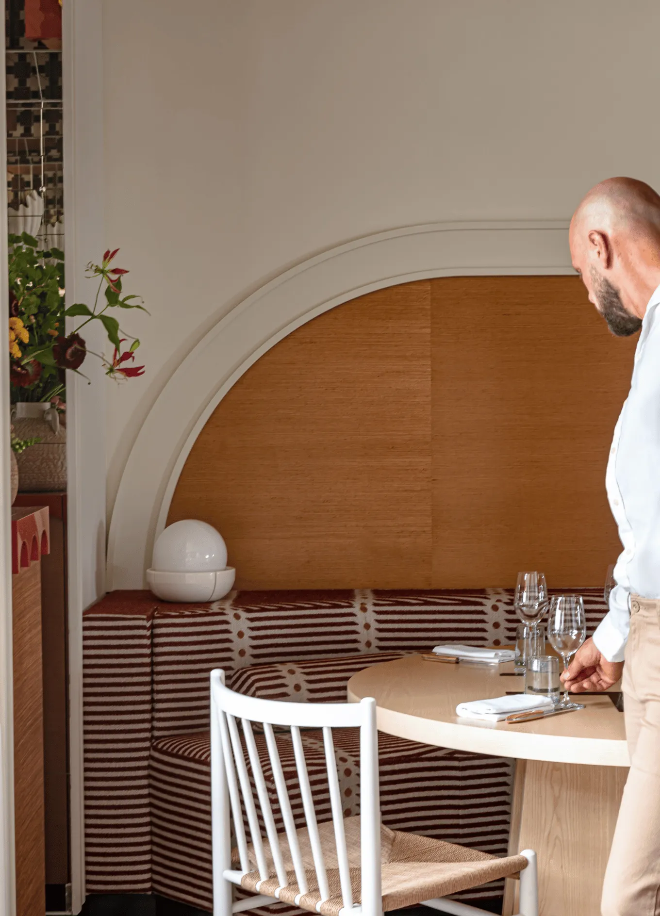 Man in white shirt setting a table in a restaurant with patterned booth seating and a wooden round table.