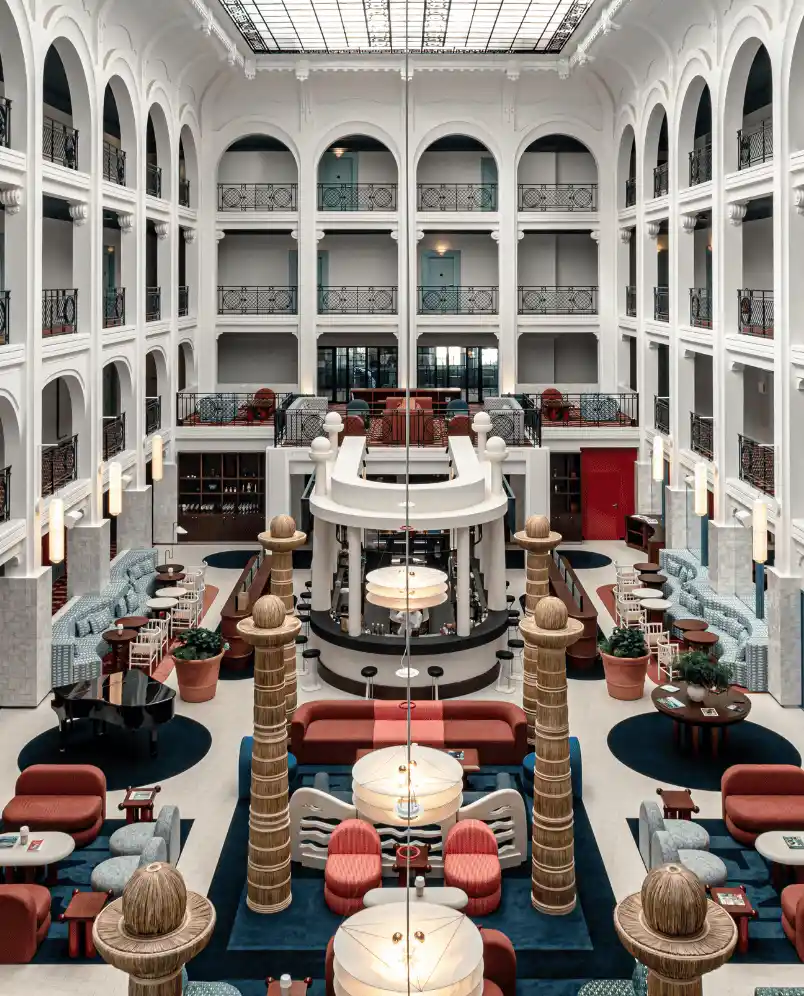 Spacious hotel atrium with white arched balconies, chic seating areas, wooden decorative columns, and large hanging lights under a glass ceiling.