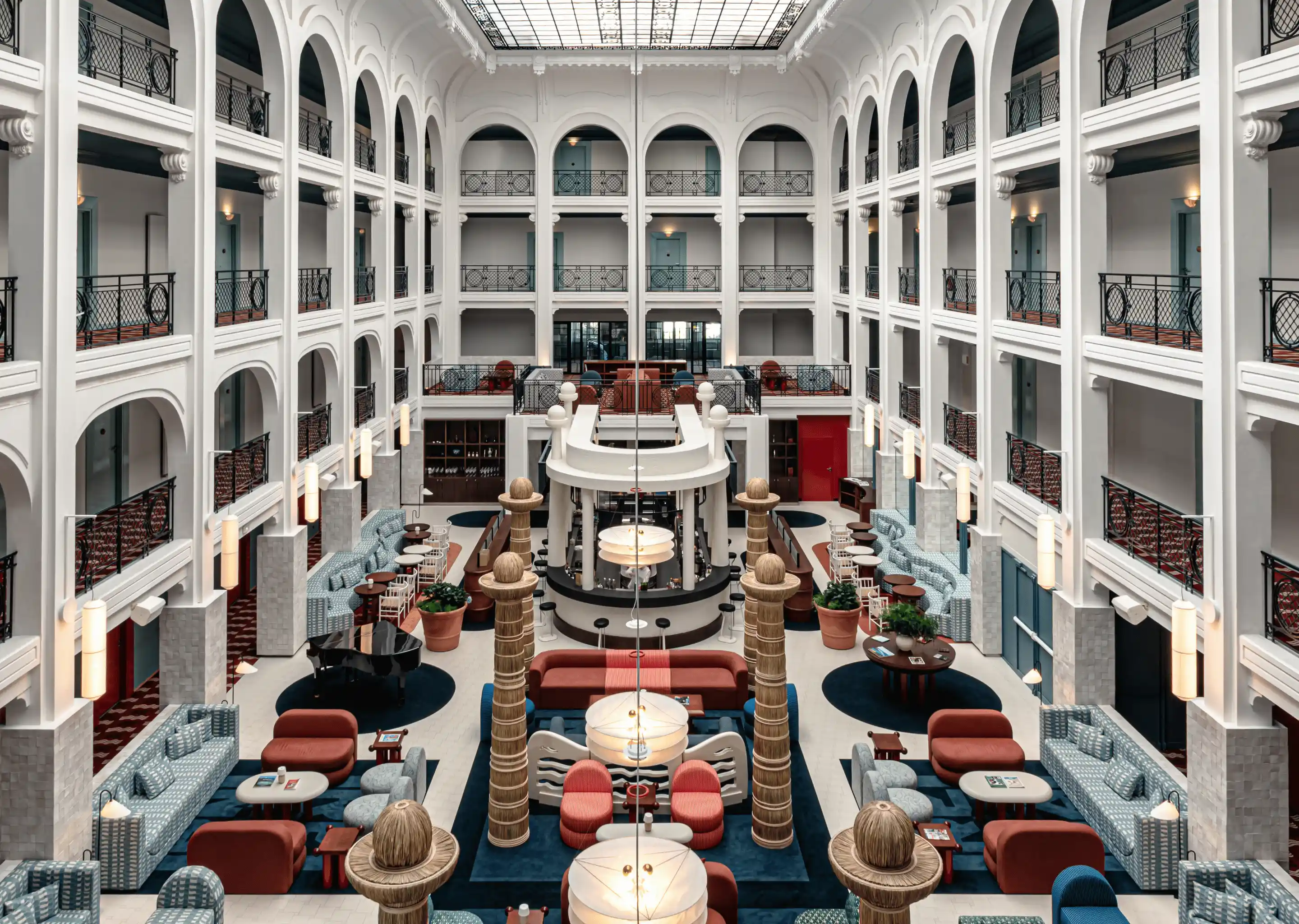 Large hotel atrium with white archways, multiple seating areas with red and blue sofas, potted plants, a black grand piano, and a glass ceiling.
