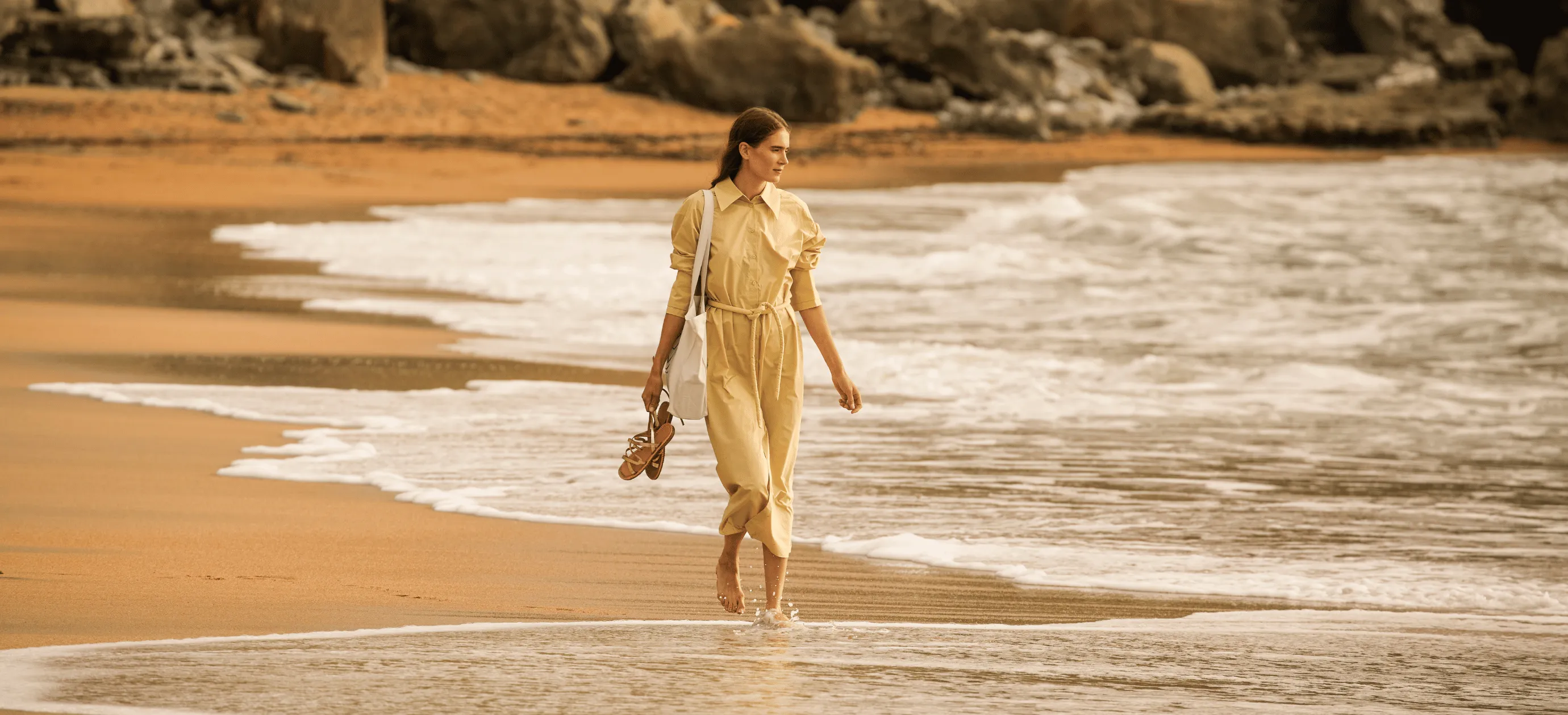 Woman in beige dress walking barefoot along a sandy beach near gentle ocean waves, carrying sandals and a shoulder bag.
