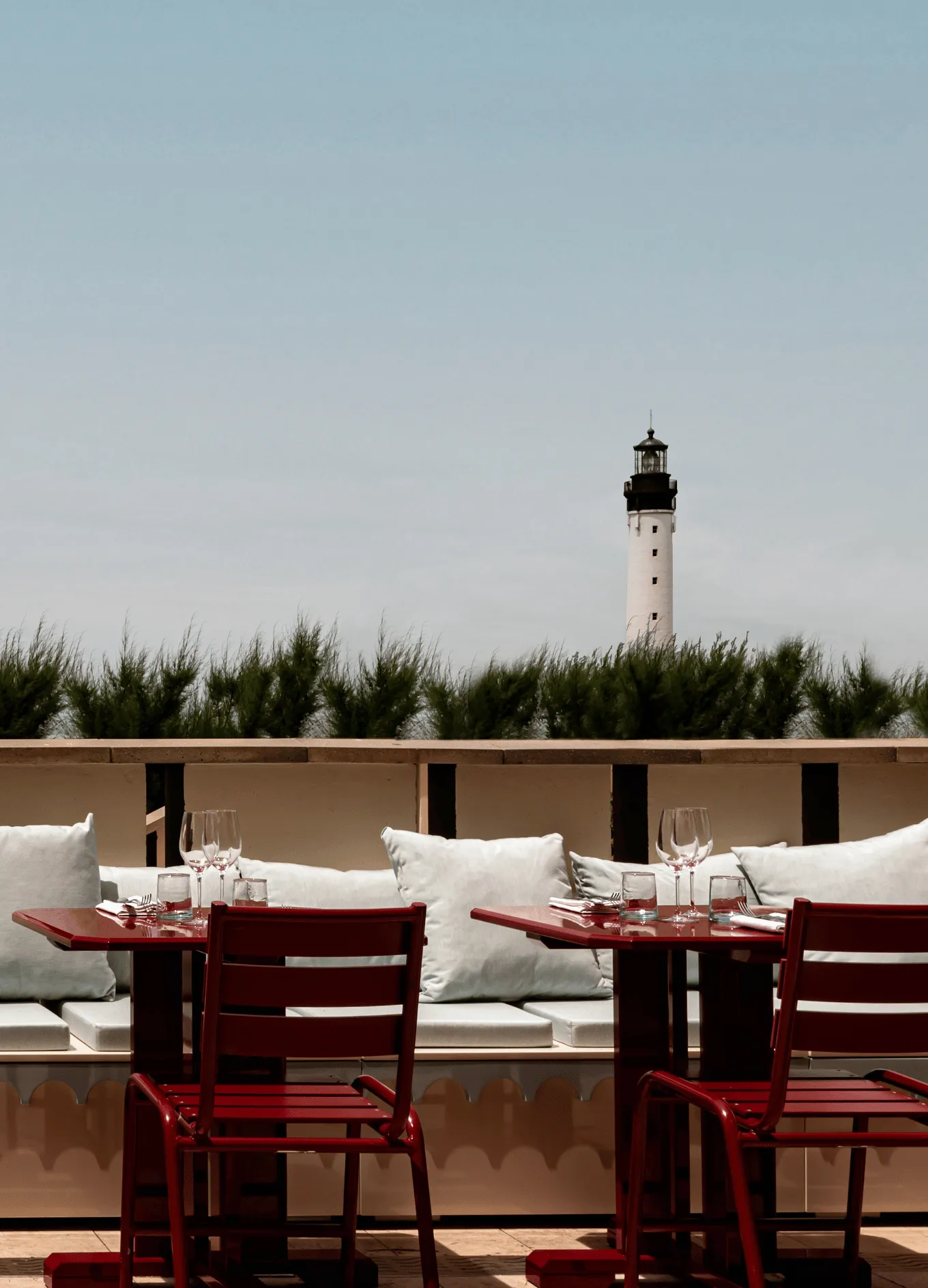 Outdoor seating area with red chairs and tables set with glasses in front of a cushioned bench, with a lighthouse visible in the background.
