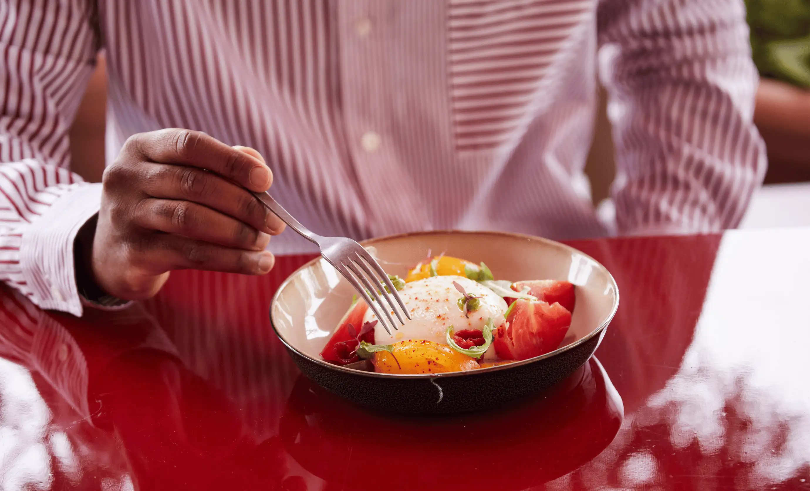 Person holding a fork over a bowl with two sunny-side-up eggs, sliced tomatoes, and greens on a red table.