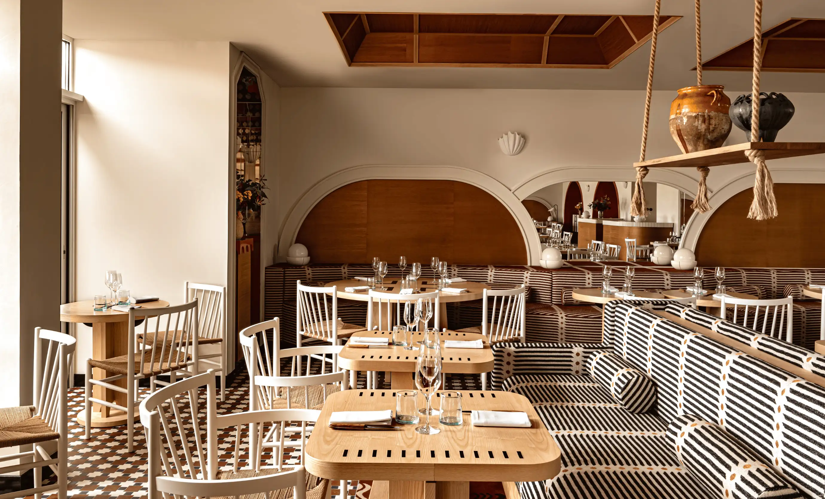 Elegant restaurant interior with wooden tables, white chairs, and striped cushioned seating, featuring a hanging shelf with pottery.