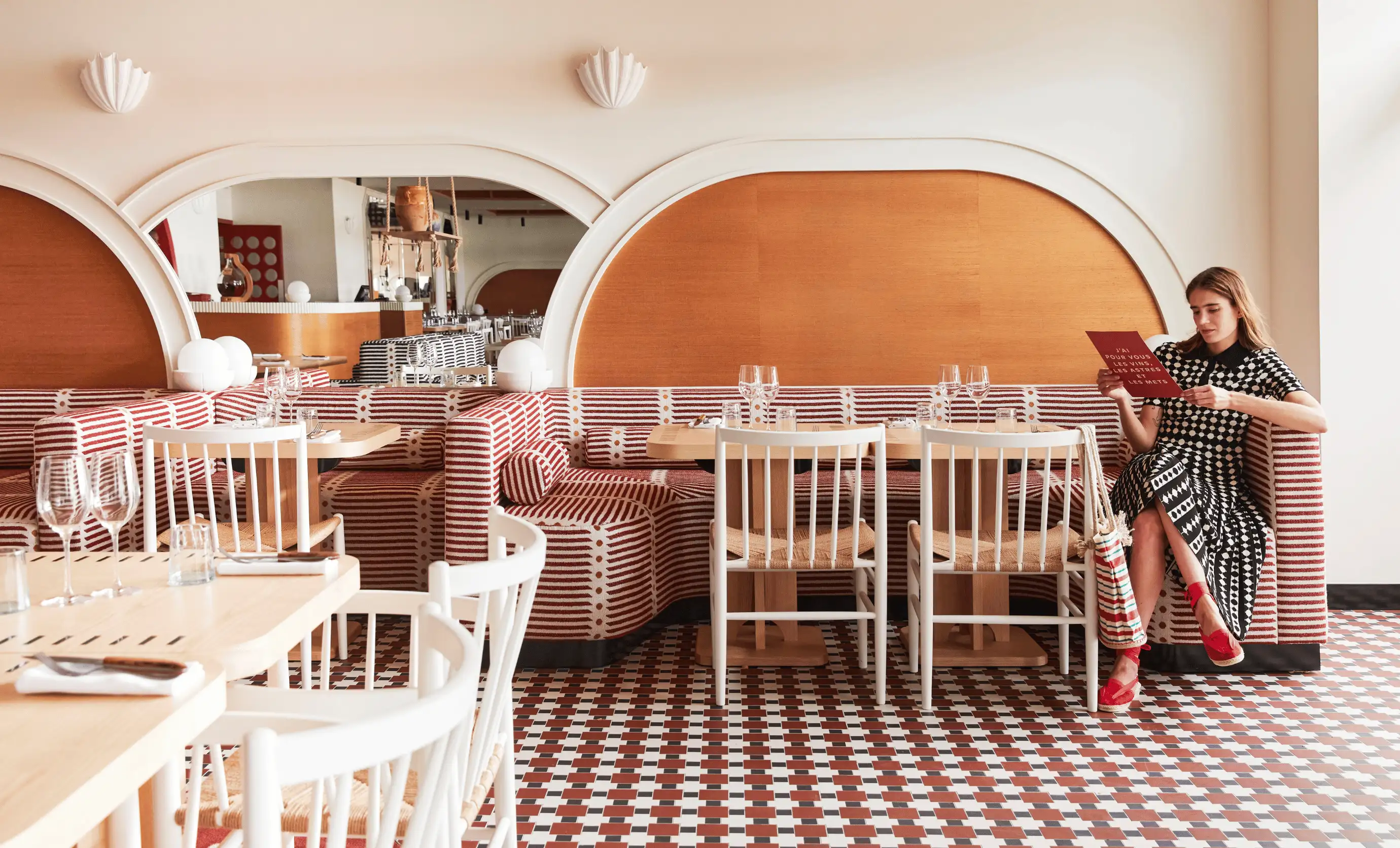 Woman in a patterned dress sitting on a striped banquette in a stylish café with checkered tile floor, reading a menu.