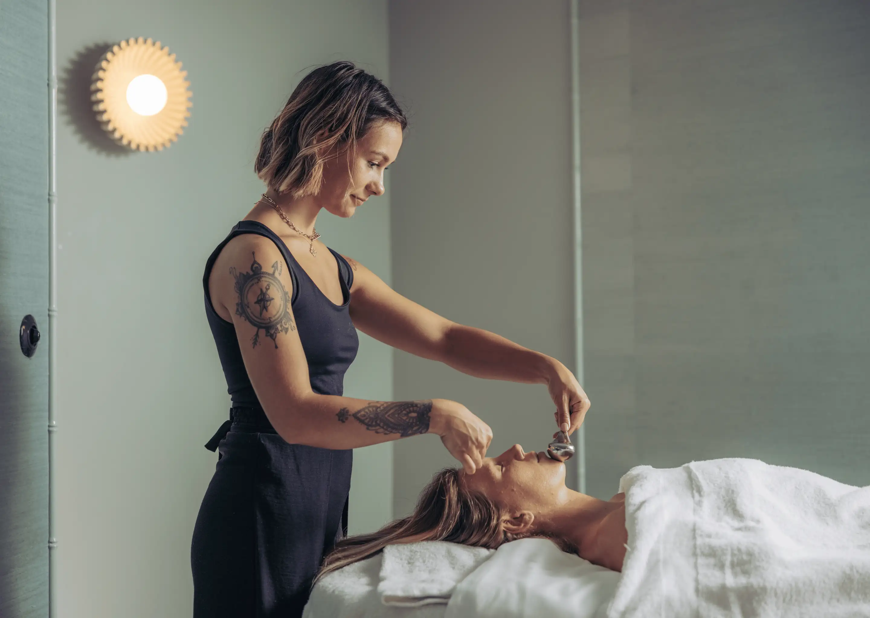 Tattooed woman giving a facial treatment with a metal tool to a relaxed woman lying on a spa bed covered with a white towel.
