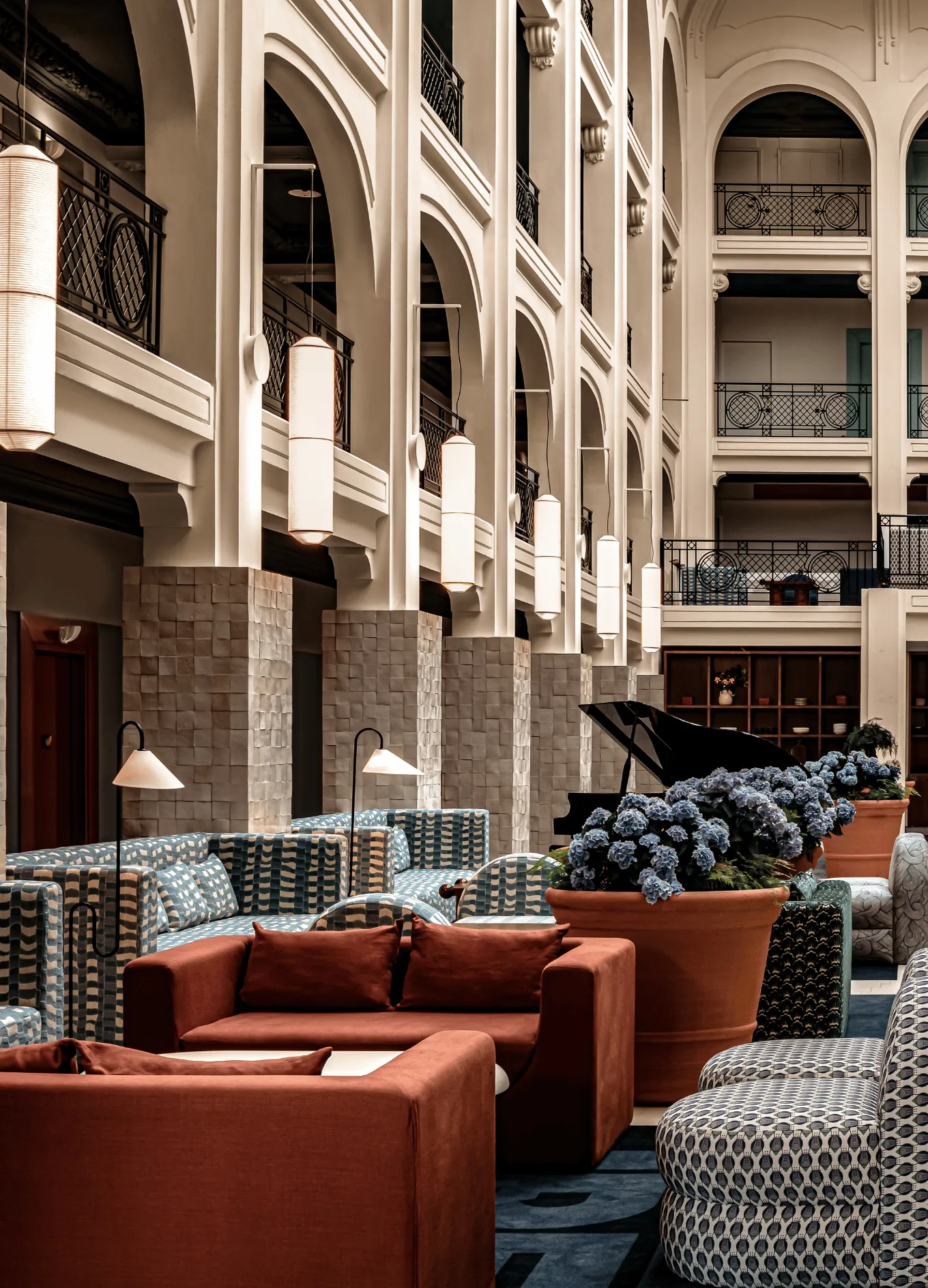 Elegant hotel lounge with patterned armchairs, rust-colored sofas, large potted blue flowers, hanging lanterns, and a black grand piano under high arched ceilings.