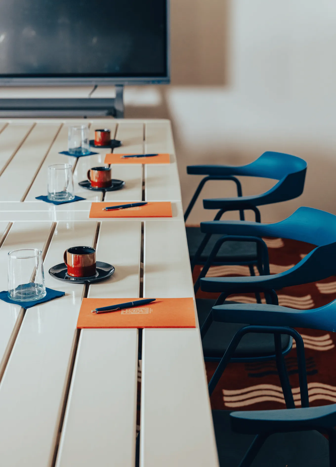 Conference table set with orange folders, pens, clear glasses on blue coasters, and brown coffee cups with saucers alongside blue chairs.