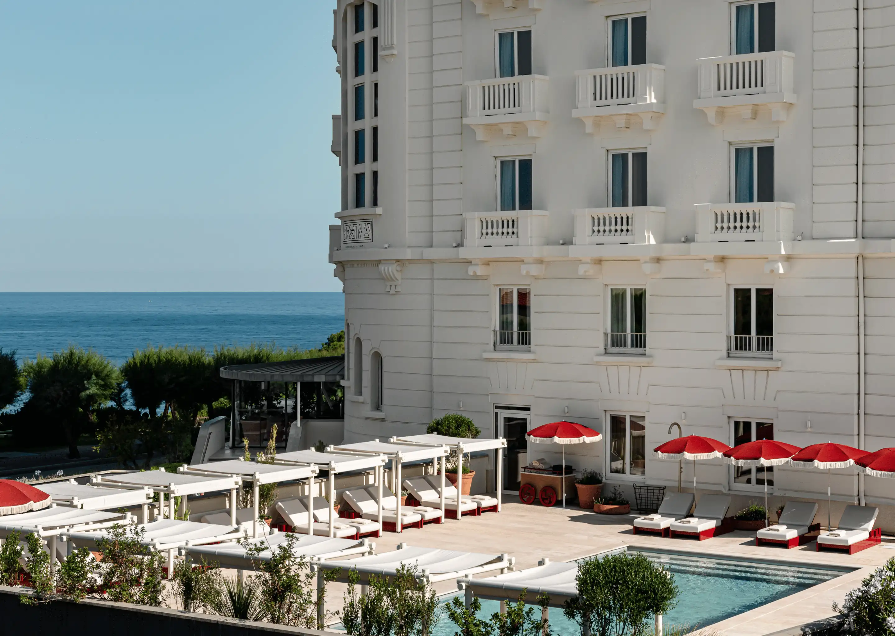 Outdoor pool area with white lounge chairs under white and red umbrellas next to a white hotel building overlooking the sea.
