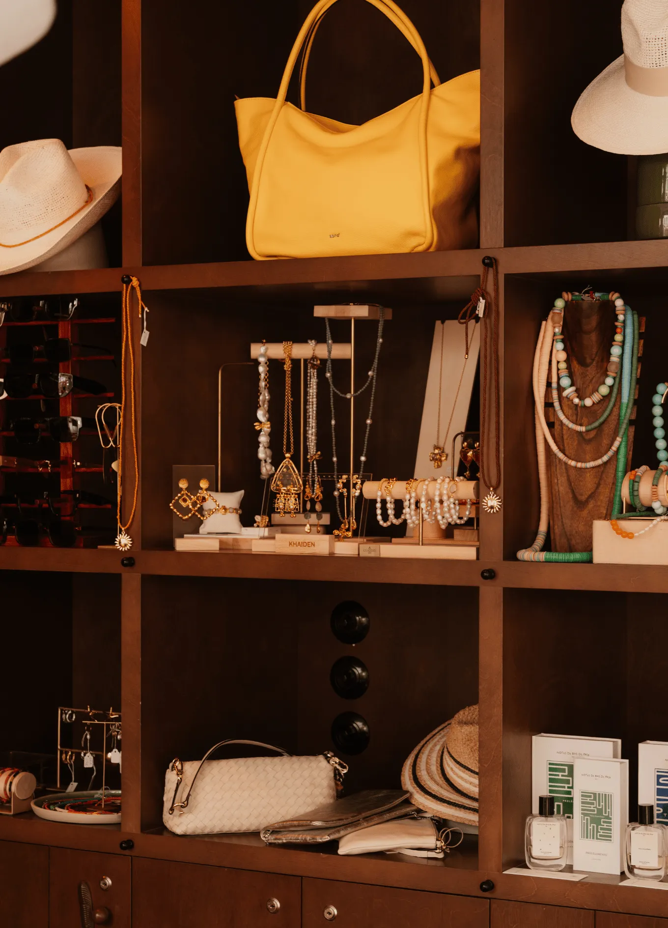 Wooden cubby shelves displaying yellow handbag, hats, sunglasses, pearl and gold jewelry, beaded necklaces, and perfume bottles.