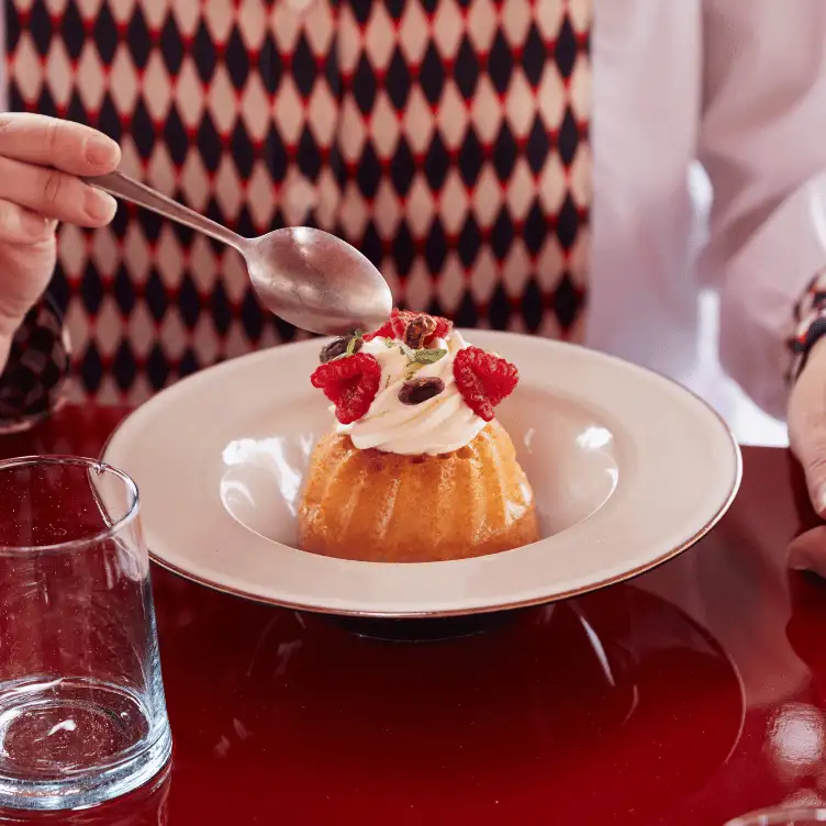Person about to eat a dessert topped with whipped cream, raspberries, and nuts, on a white plate placed on a red table.