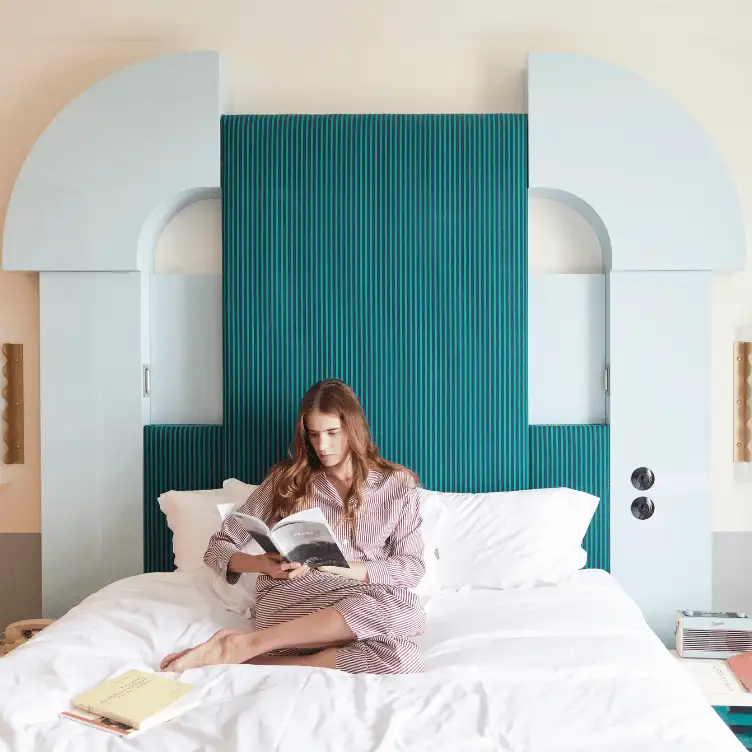 Woman sitting cross-legged on a bed with white sheets, reading a book, against a blue tufted headboard with modern light blue architectural side panels.