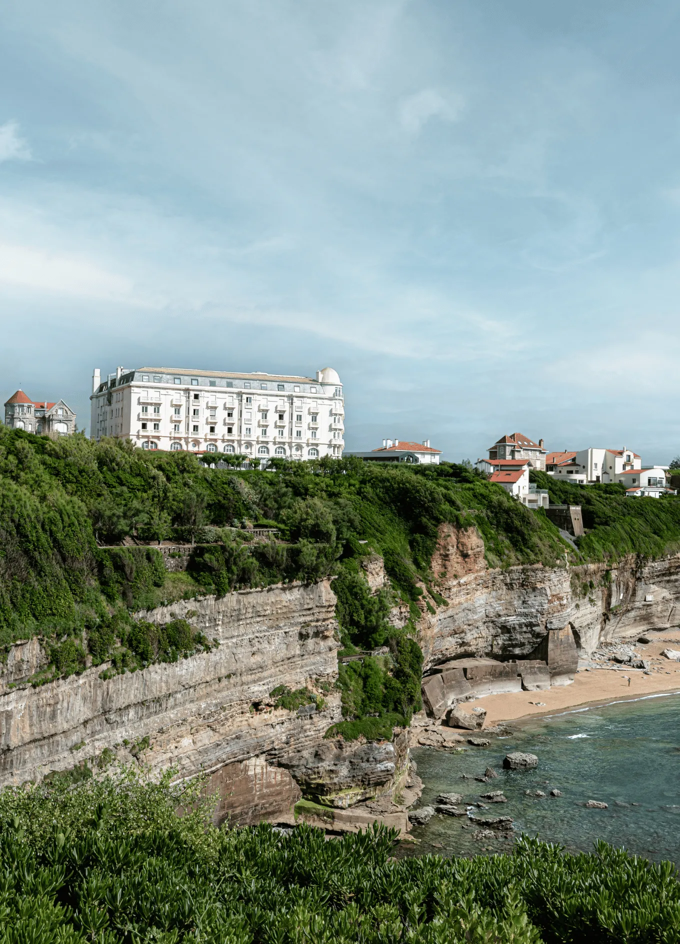 Historic white hotel and houses on a green cliff overlooking a rocky beach and calm ocean under a cloudy sky.