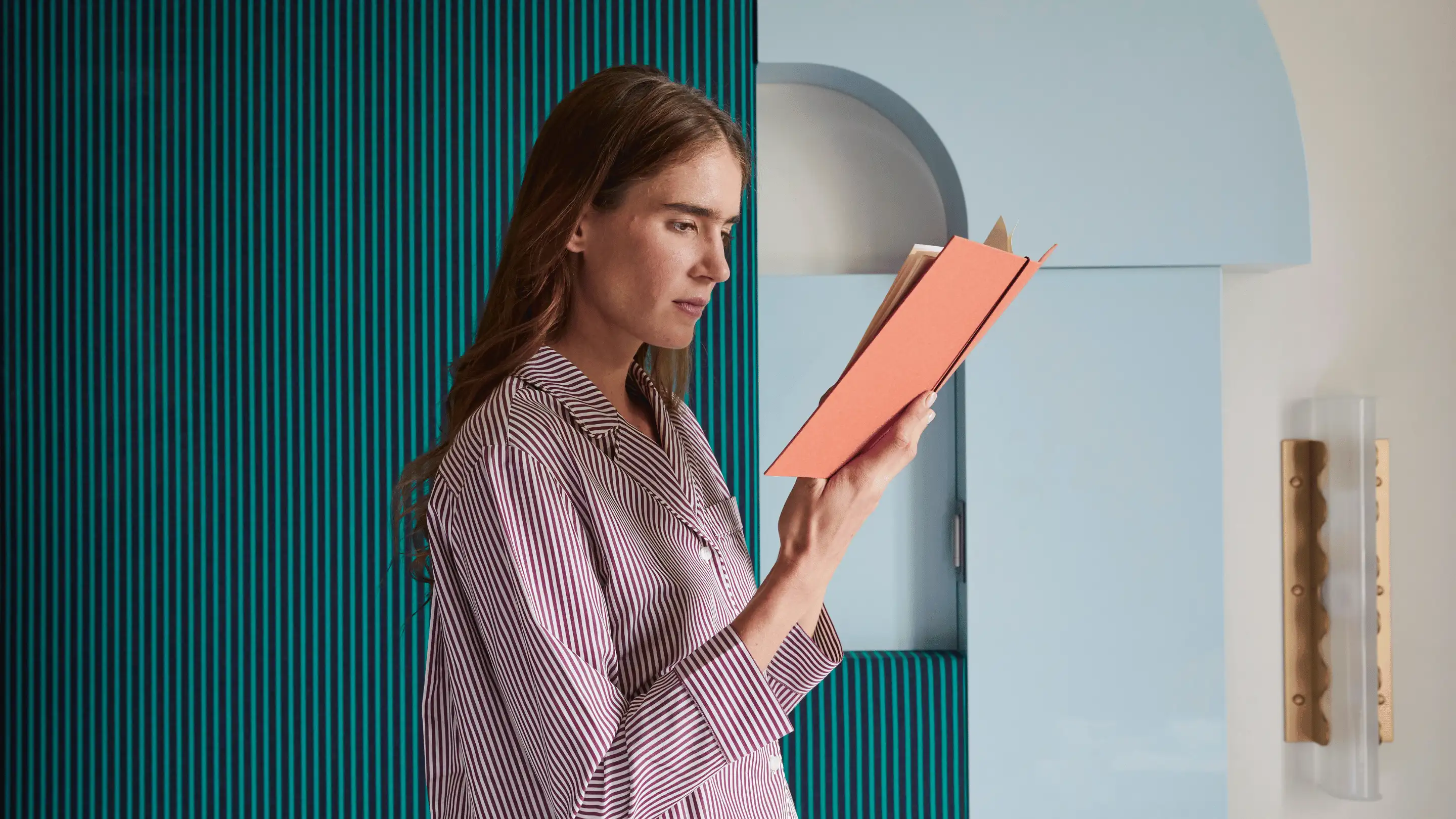 Woman in striped pajamas reading a book with an orange cover in a room with blue and teal striped wall and a blue cabinet.