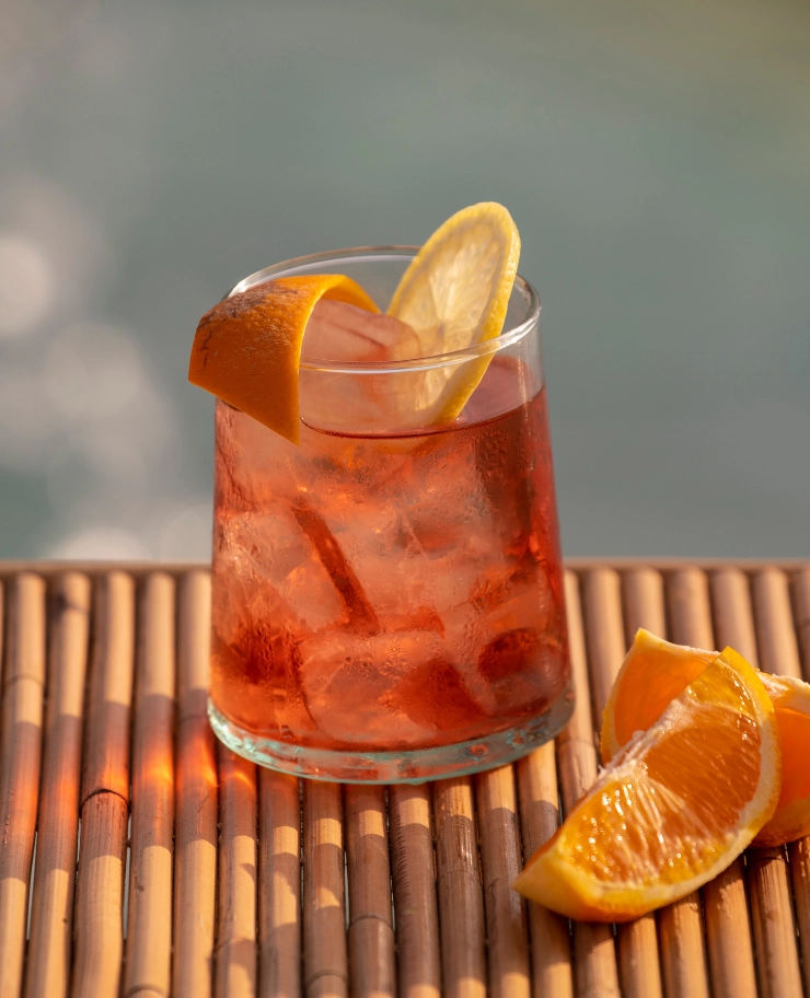 Glass of iced red cocktail garnished with a lemon slice and an orange peel on a bamboo surface with orange wedges beside it.