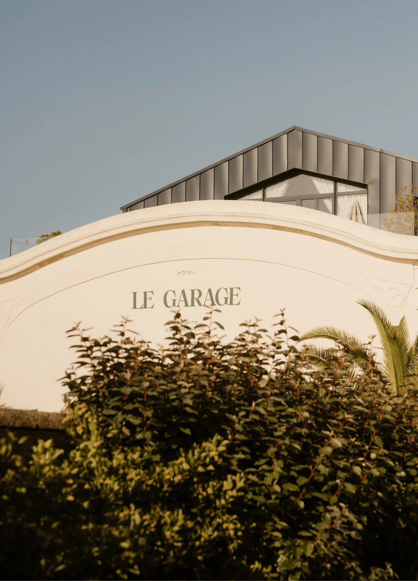 Facade of Hôtel Le Garage with beige arch, modern upper floor, and green shrubbery in the foreground.
