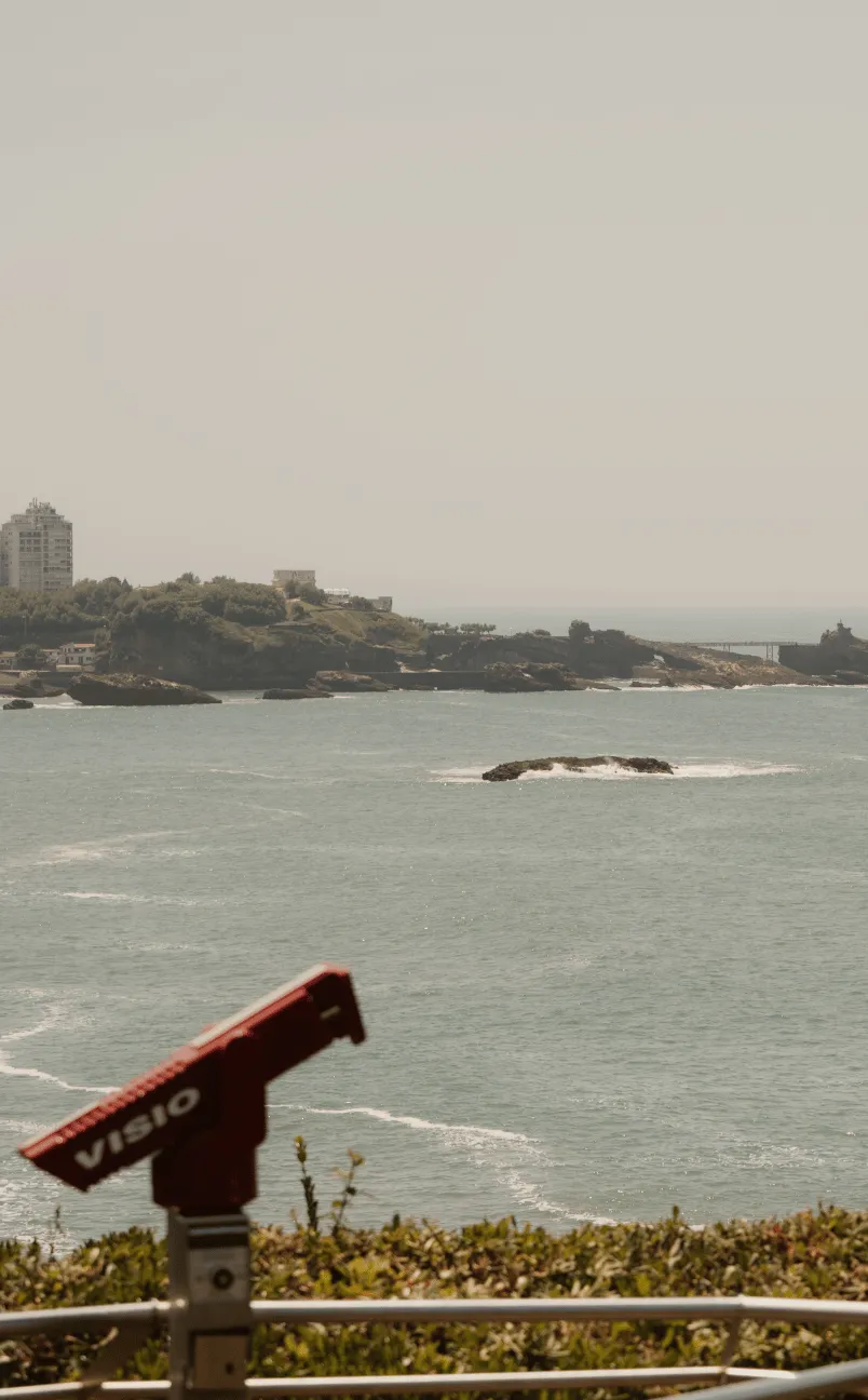 Binoculares rojos que funcionan con monedas y vistas a un océano tranquilo con una costa rocosa y edificios distantes bajo un cielo brumoso.