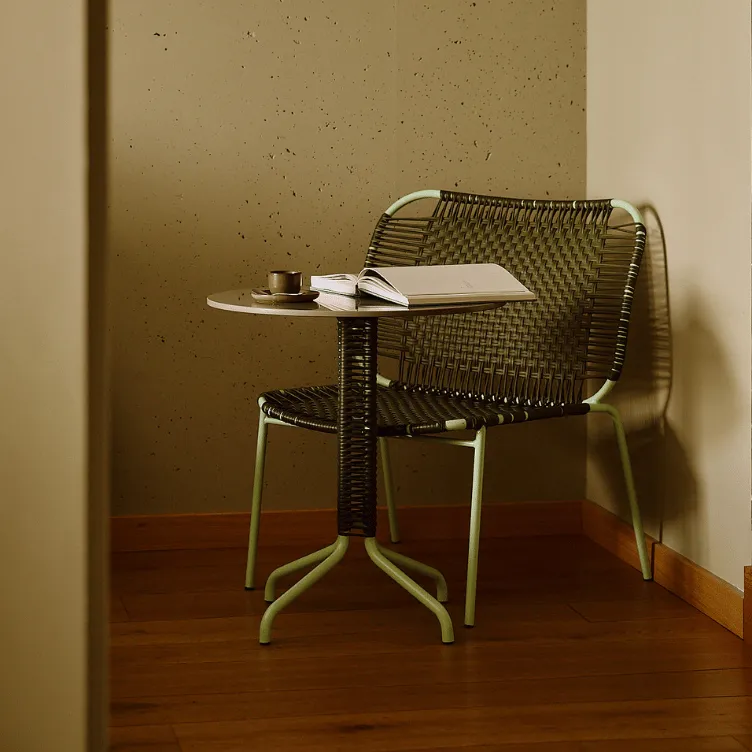 Woven chair with green metal legs next to a small round table holding an open book and a small cup on a saucer in a corner with textured beige walls.
