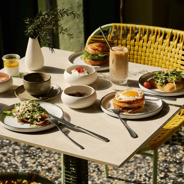 Sunlit table with brunch dishes including a sandwich, iced coffee, pancakes topped with a fried egg, toast with greens, and small bowls of yogurt and granola.