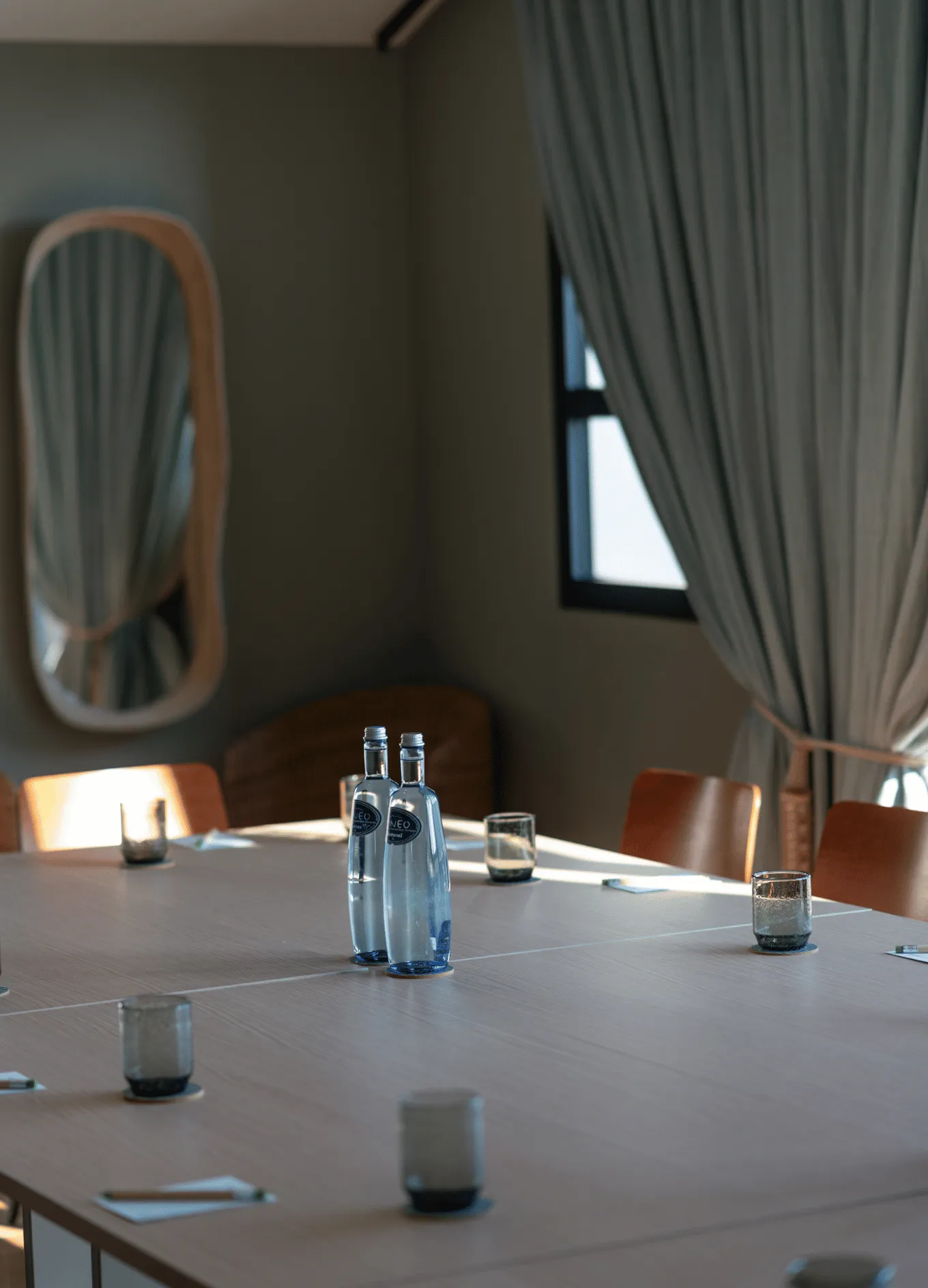 Moderna sala conferenze con tavolo in legno, sedie marroni, due bottiglie d'acqua, bicchieri vuoti e luce naturale da una finestra con tende.