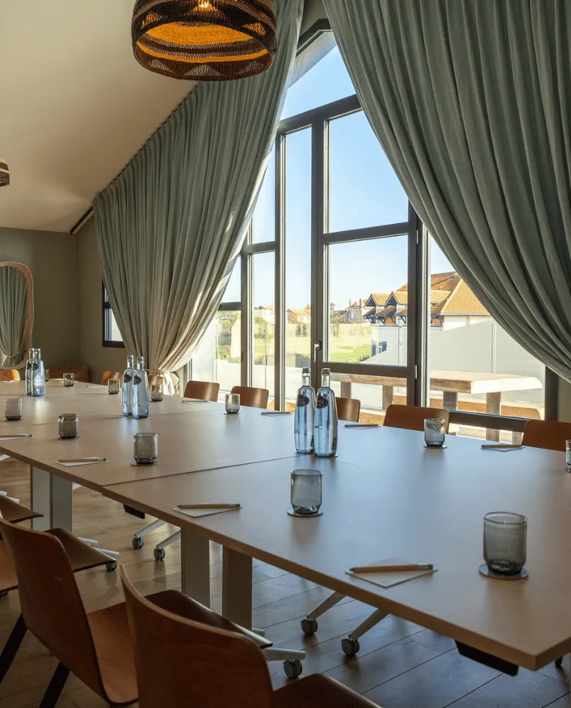 Moderna sala conferenze con lunghi tavoli bianchi, sedie marroni, bottiglie d'acqua, bicchieri e grandi finestre con tende verdi.
