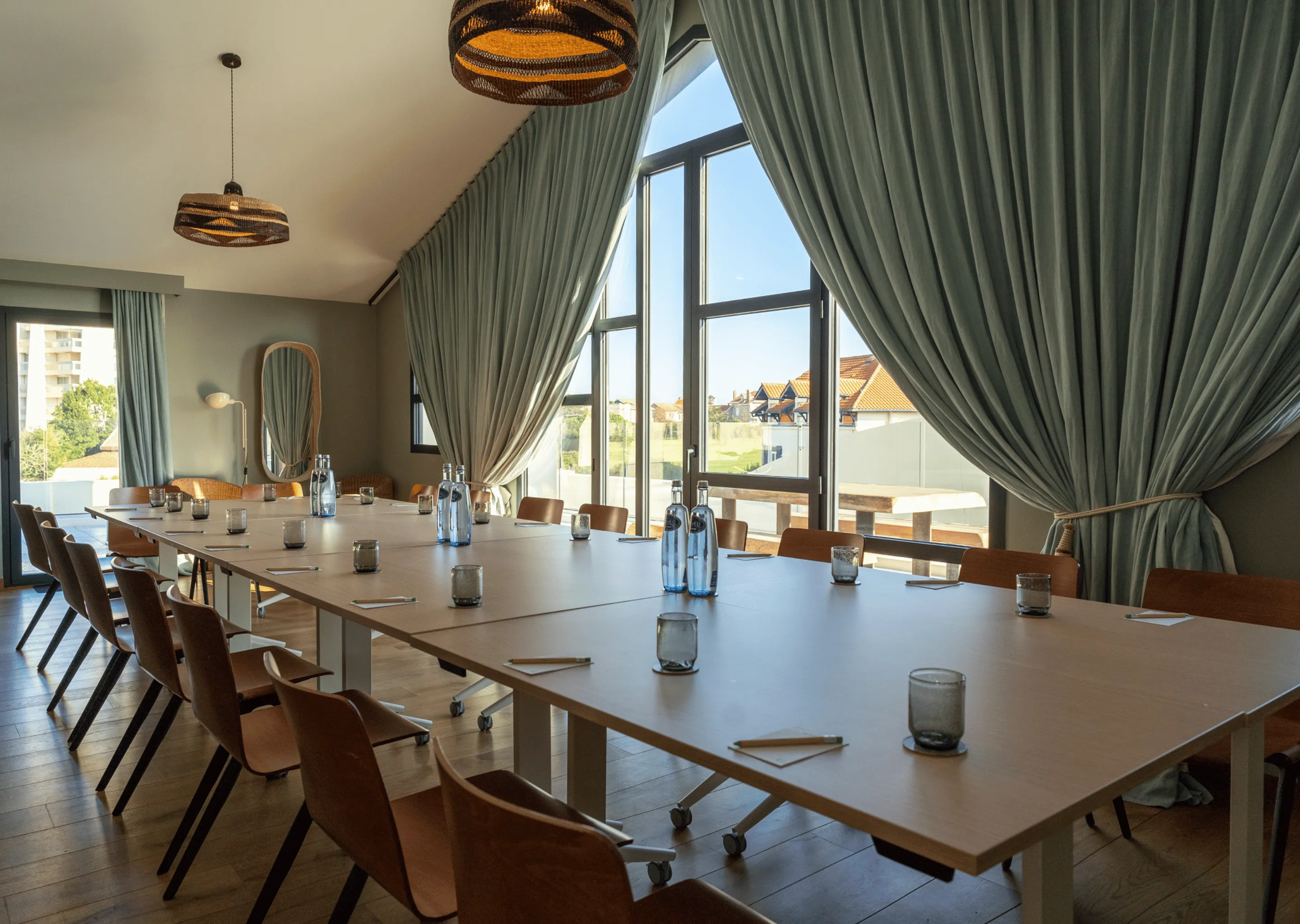 Sala conferenze con un grande tavolo rettangolare circondato da sedie in legno, con bicchieri, taccuini e bottiglie d'acqua, illuminata dalla luce naturale proveniente da alte finestre con tende.