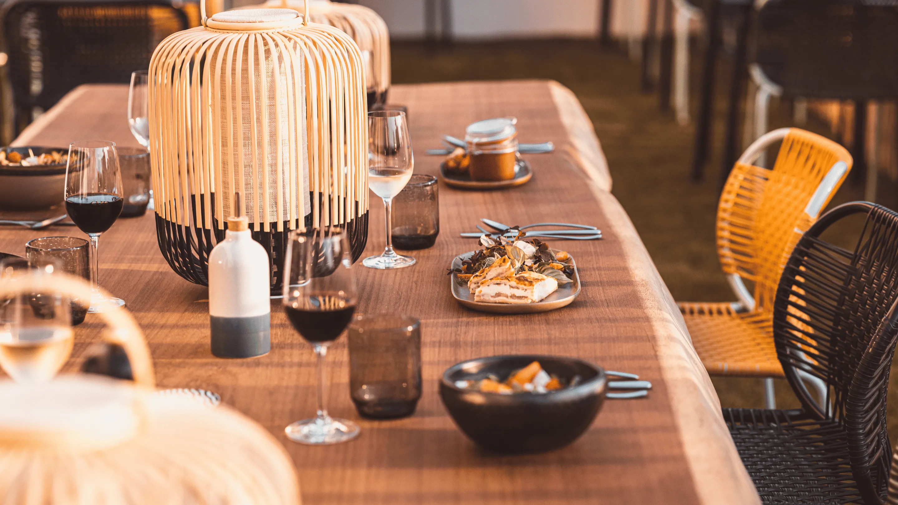 Lungo tavolo da pranzo in legno con bicchieri da vino, lanterne decorative, piatti con cibo e sedie intrecciate con luci calde.